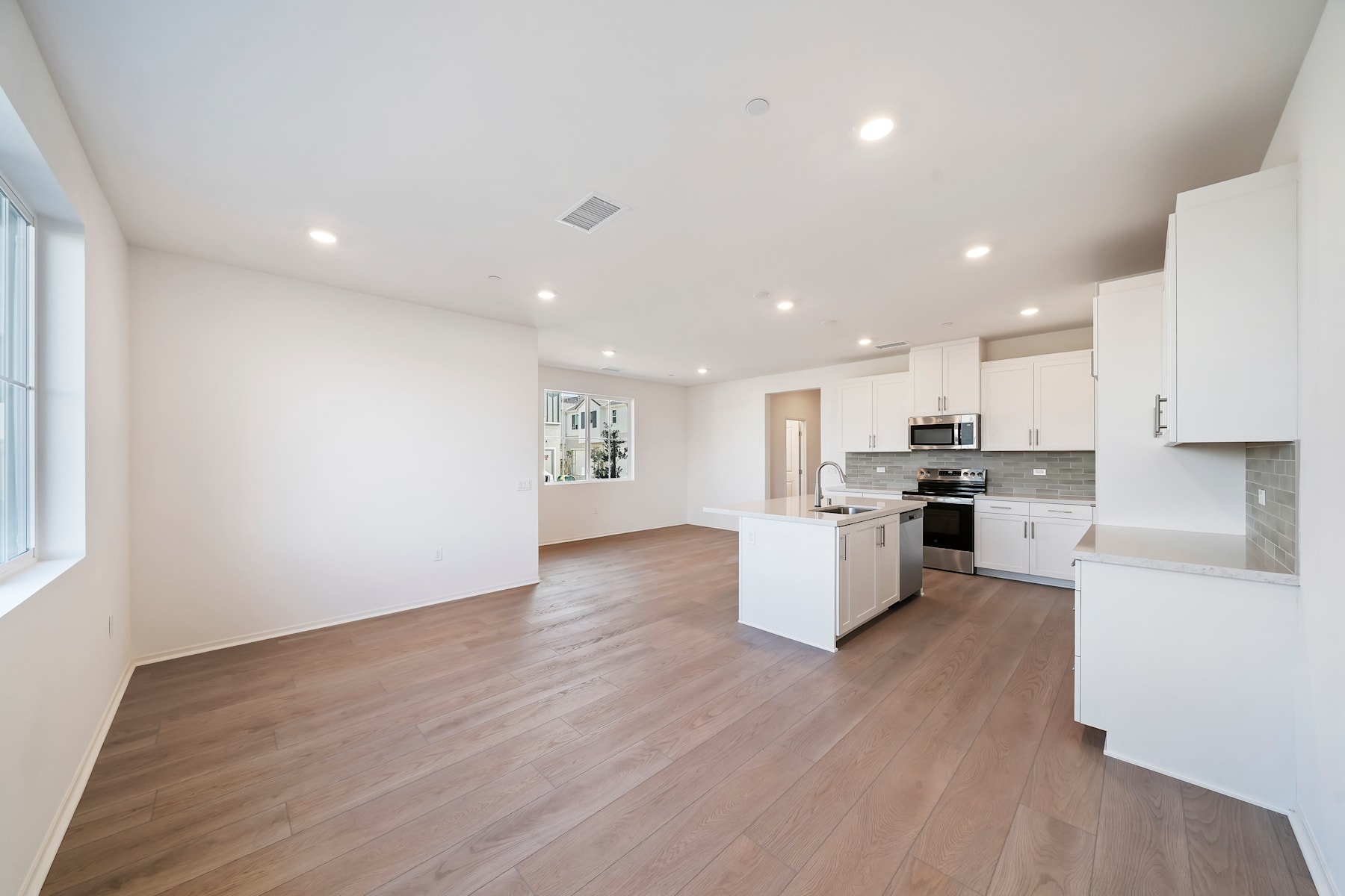 A spacious and modern kitchen with white cabinets, stainless steel appliances, and hardwood floors, leading into an open living area with a large window.