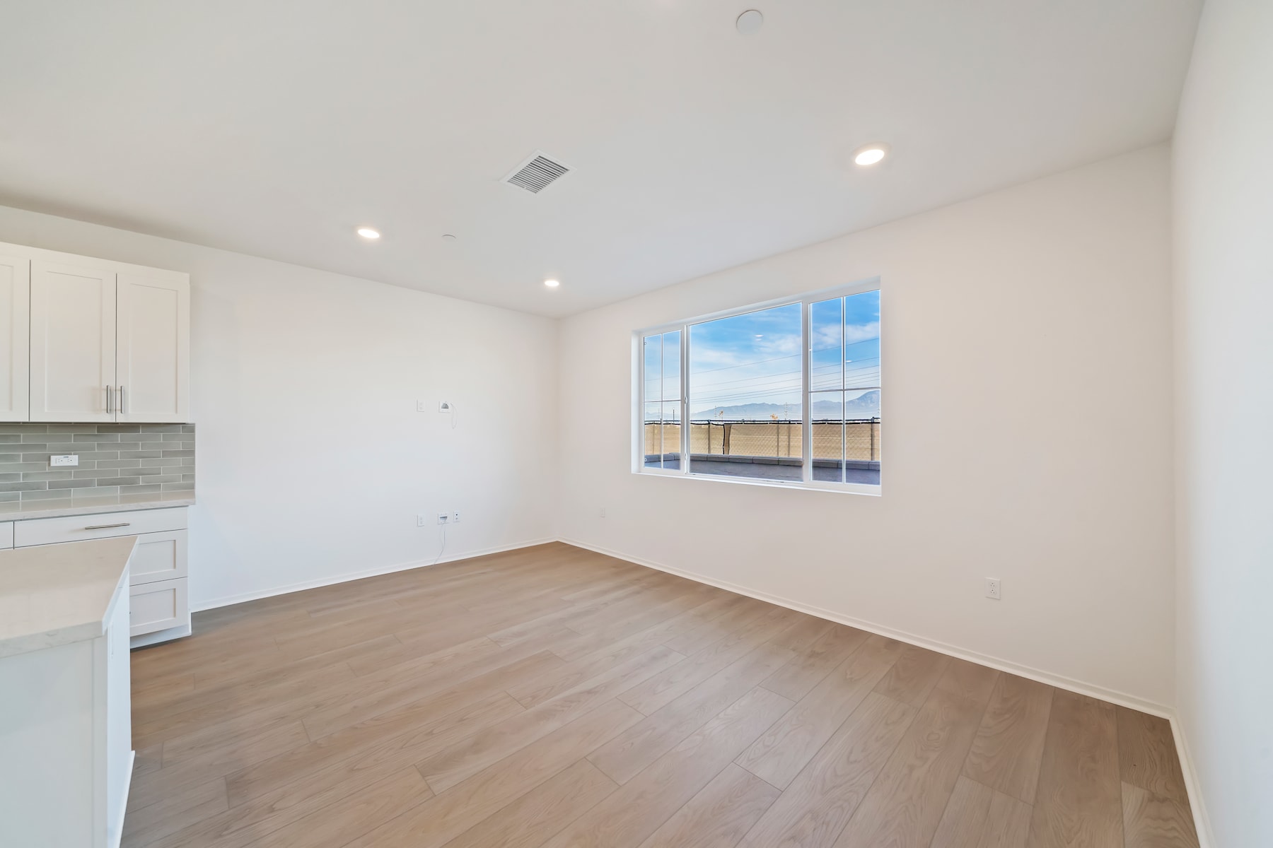 A bright and spacious room with a large window overlooking the outdoors, featuring hardwood flooring and a kitchen area visible in the background.