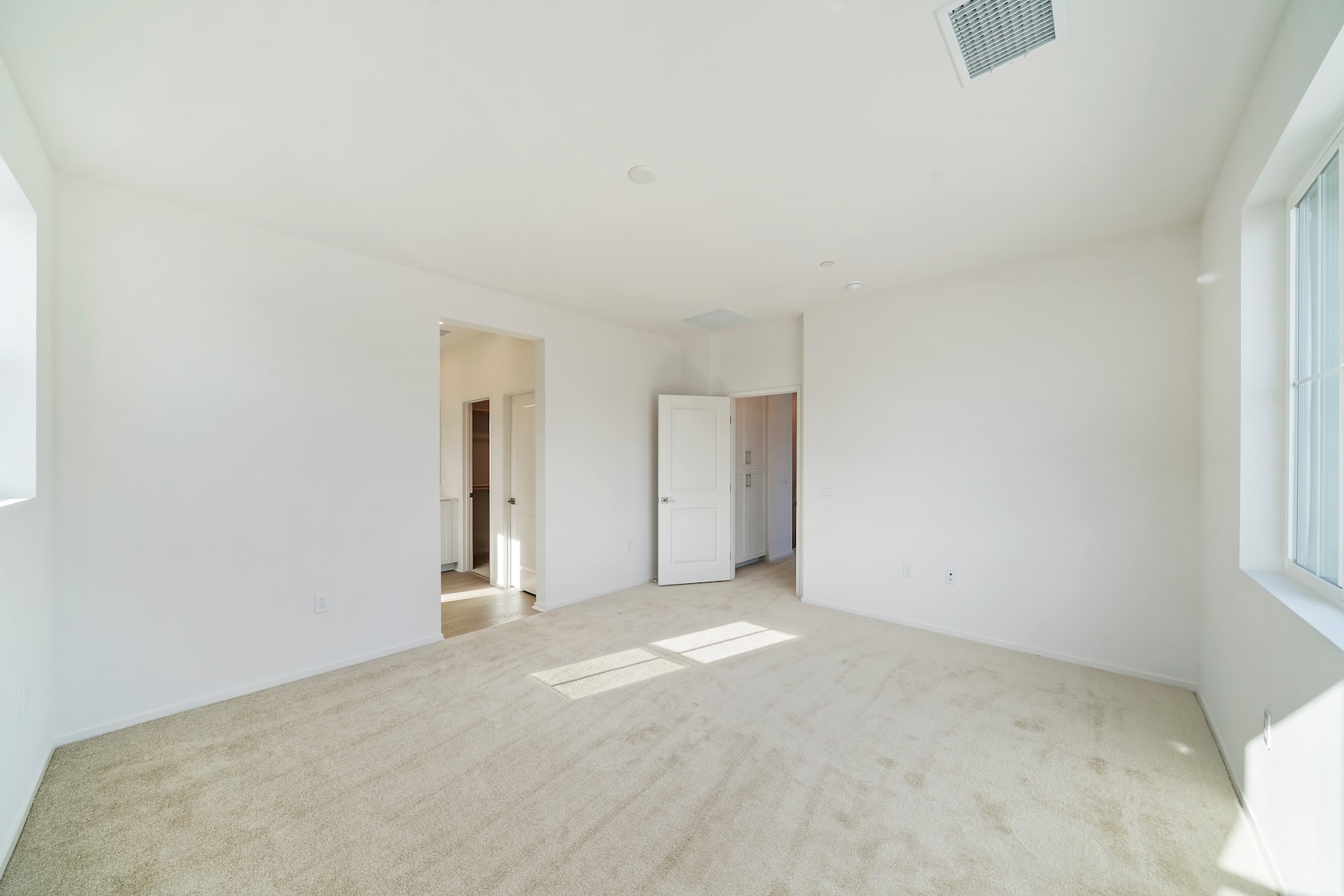 A spacious, minimalist room with white walls, a hardwood floor, and a window allowing natural light to stream in.