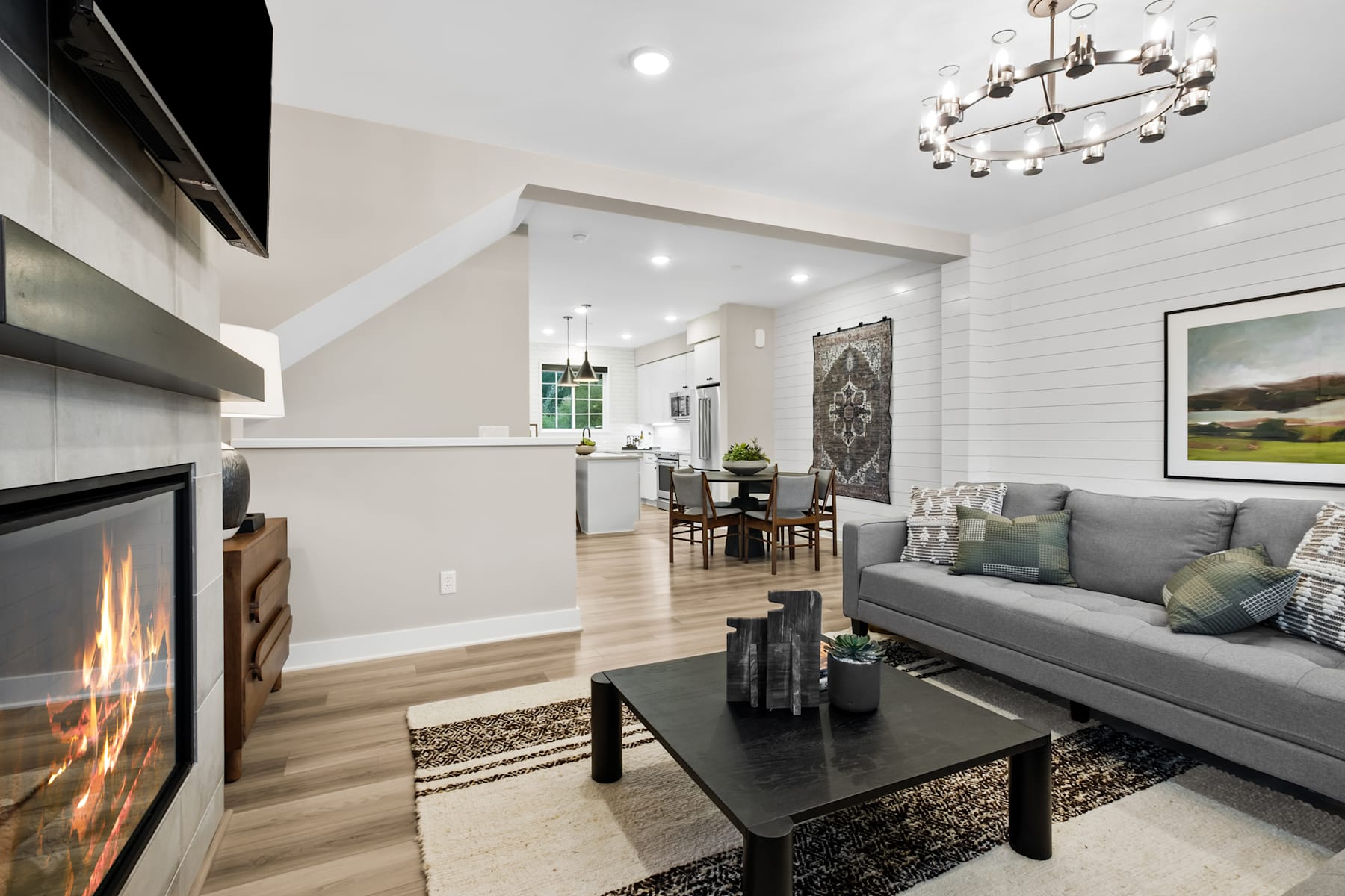 A cozy and modern living room with a fireplace, a gray sofa, a black coffee table, and a chandelier hanging from the ceiling, leading into a dining area with a wooden table and chairs.