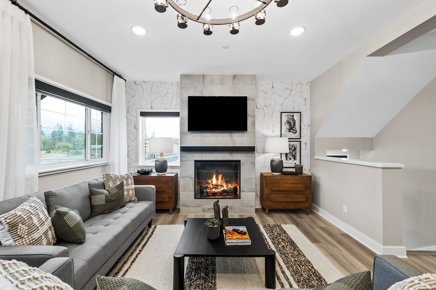 A cozy and modern living room with a fireplace, a gray sofa, wooden furniture, and a chandelier hanging from the ceiling.