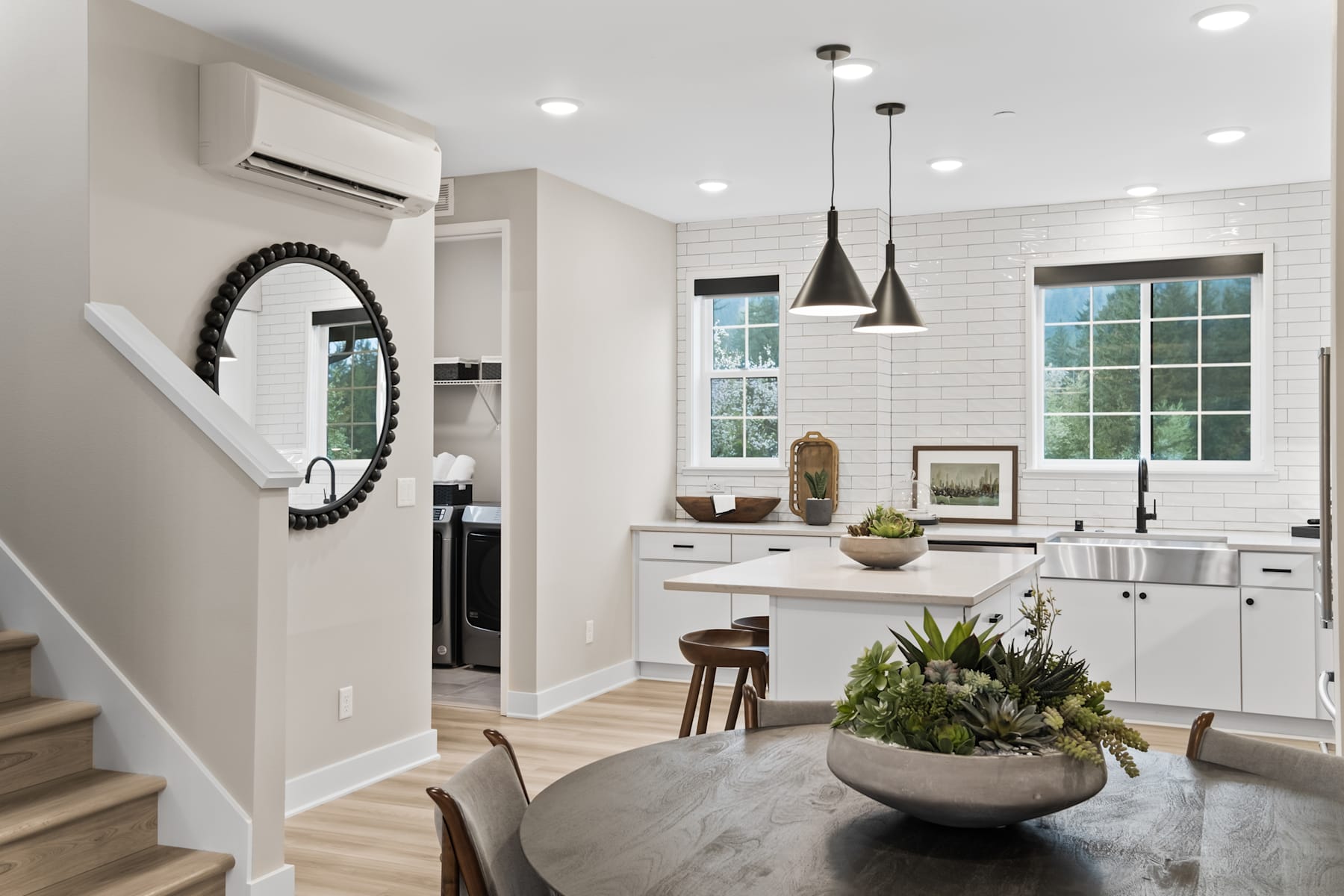 A modern, open-concept kitchen and living space with white walls, wooden floors, and a large mirror on the wall, complemented by potted plants and pendant lighting.