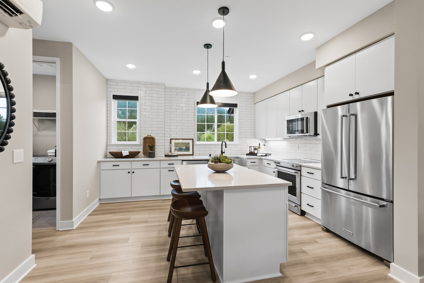 A modern, well-lit kitchen with white cabinets, stainless steel appliances, and a central island with bar stools, set against a backdrop of large windows overlooking a scenic outdoor view.