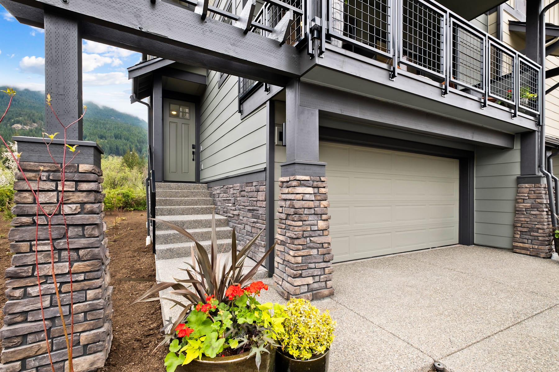 A modern, two-story house with a garage door, stone accents, and a colorful flower planter in the foreground, set against a backdrop of lush greenery and mountains.