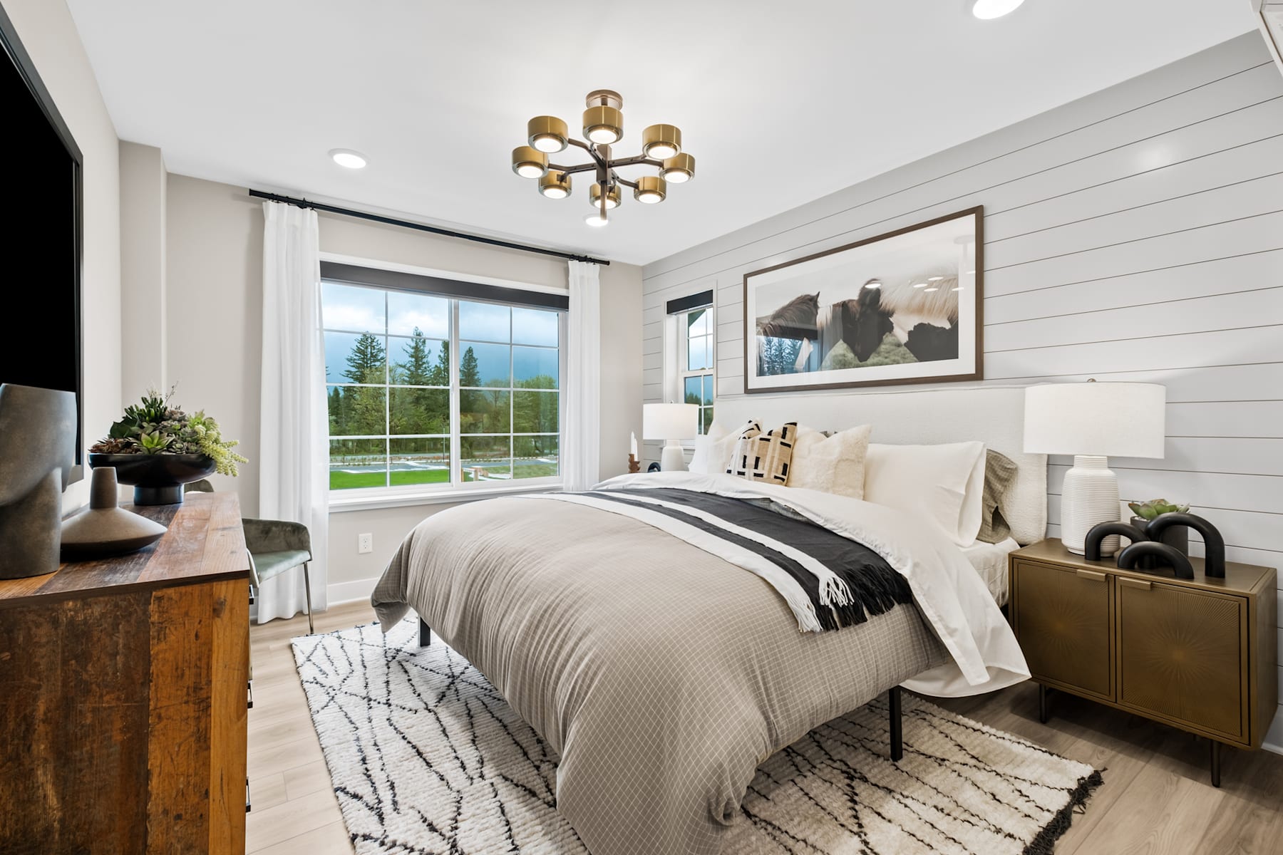 A cozy and well-appointed bedroom with a large window overlooking a scenic outdoor landscape, featuring a plush bed, a stylish chandelier, and various decorative elements that create a warm and inviting atmosphere.