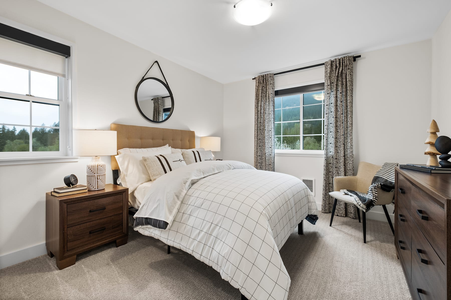 A cozy and well-furnished bedroom with a large window overlooking a scenic outdoor landscape, featuring a plaid bedspread, wooden furniture, and a circular mirror on the wall.