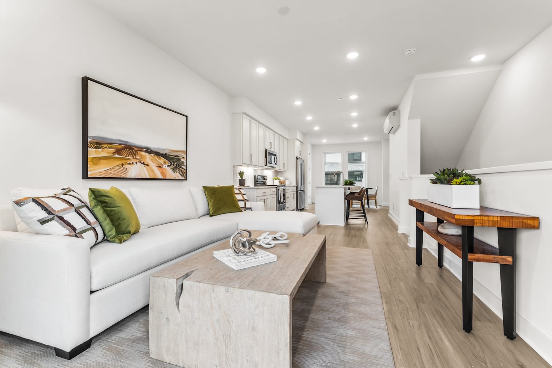 A modern and minimalist living room with a white sofa, wooden console table, and framed artwork on the wall, leading into a kitchen area with a wooden dining table.