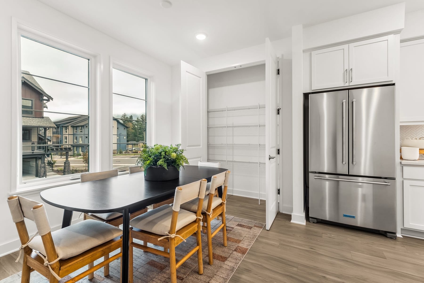 A modern, bright kitchen with a dining table, chairs, and a large stainless steel refrigerator, set against a backdrop of windows overlooking a residential neighborhood.
