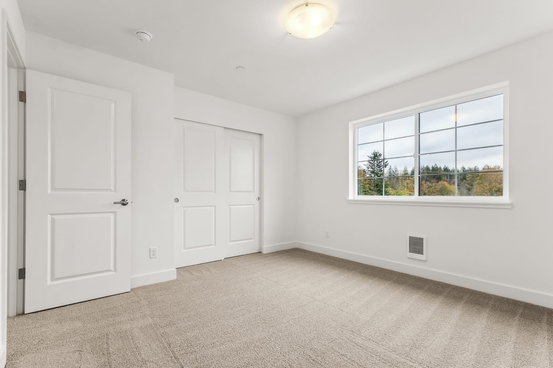 A spacious, well-lit room with white walls, a large window overlooking a forested landscape, and a hardwood floor.