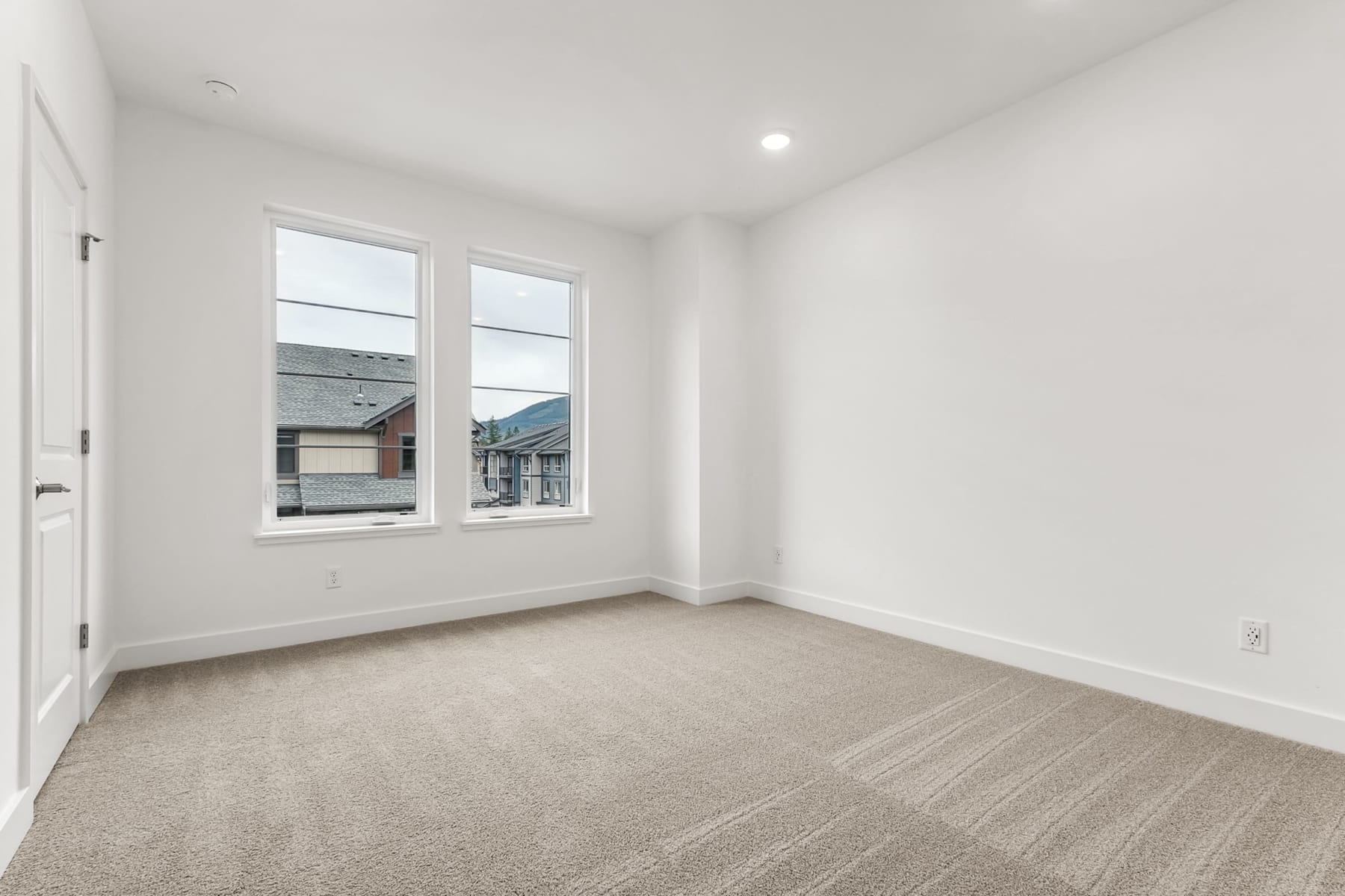A bright, empty room with white walls, a carpeted floor, and two large windows overlooking a residential neighborhood in the background.