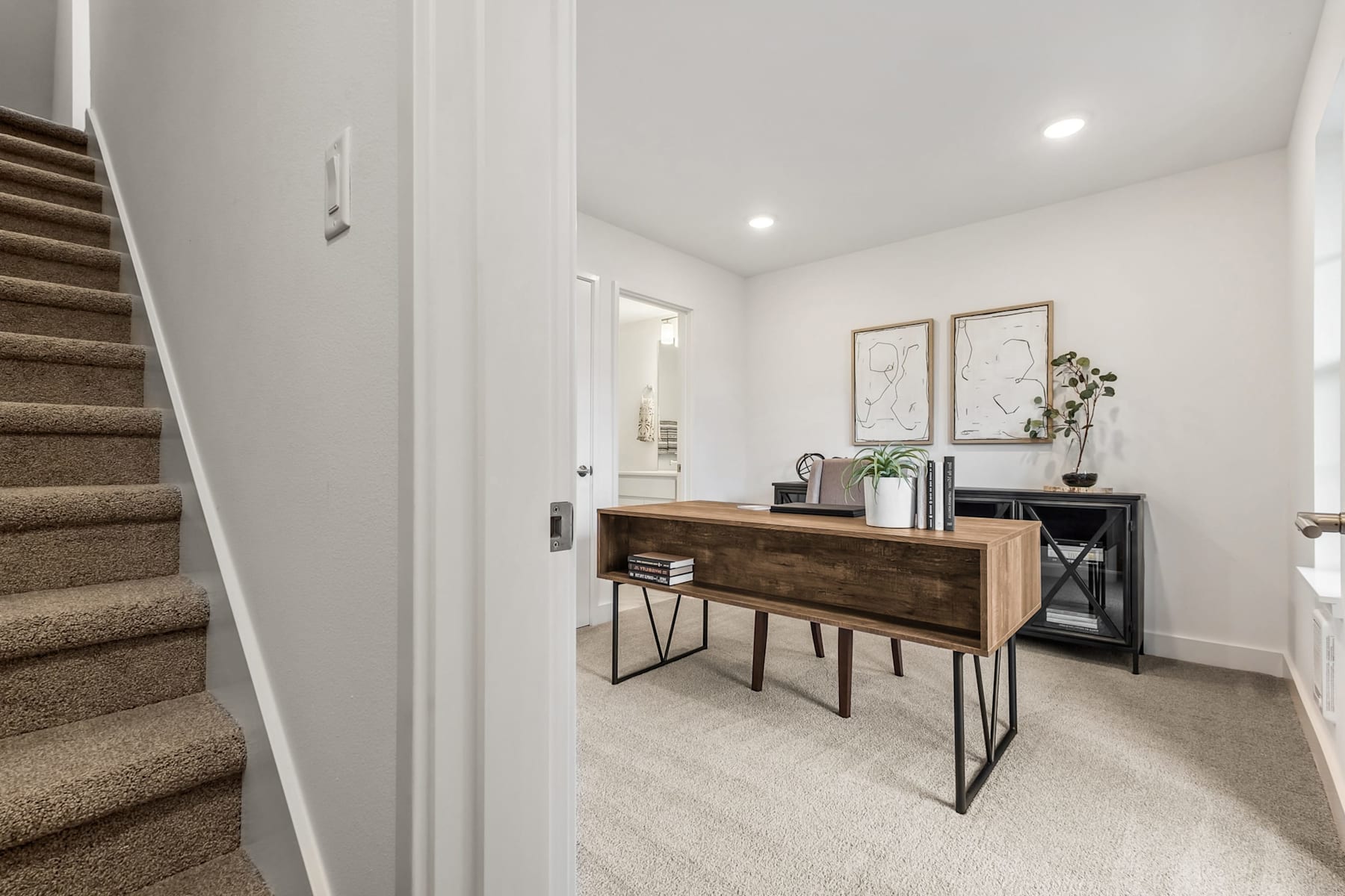 A modern and minimalist entryway with a wooden console table, decorative plants, and framed artwork on the walls, leading up to a carpeted staircase.