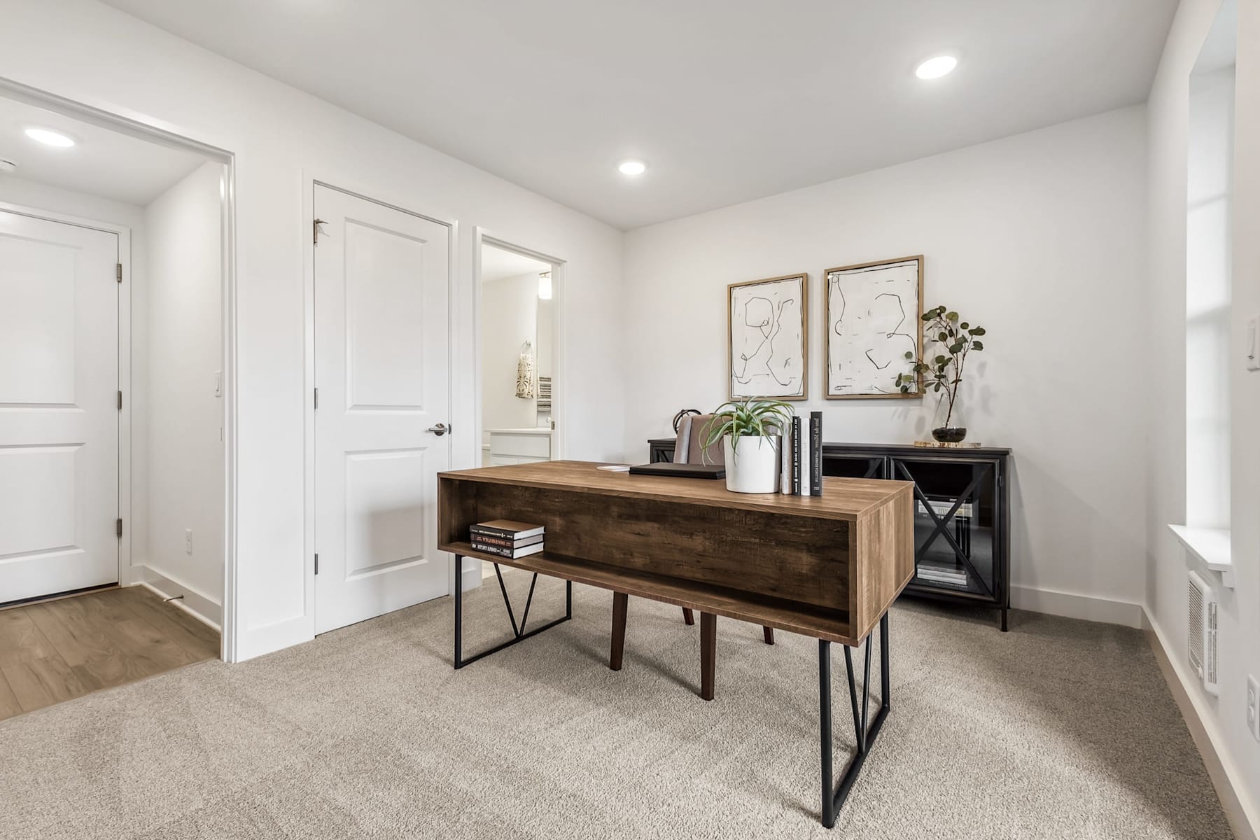 A modern and minimalist entryway with a wooden console table, framed artwork, and a potted plant, set against a neutral-toned background with white walls and doors.