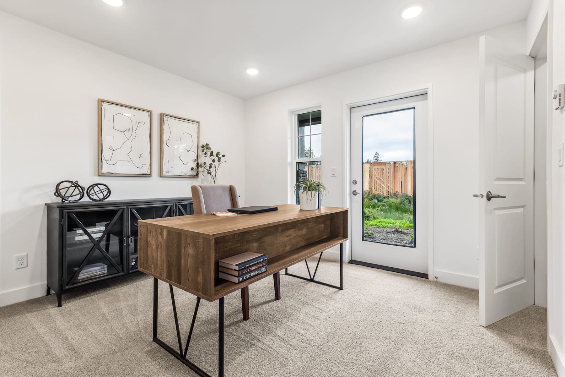 A modern and minimalist home office space with a wooden desk, black shelving unit, and framed artwork on the walls, overlooking a backyard through a glass door.
