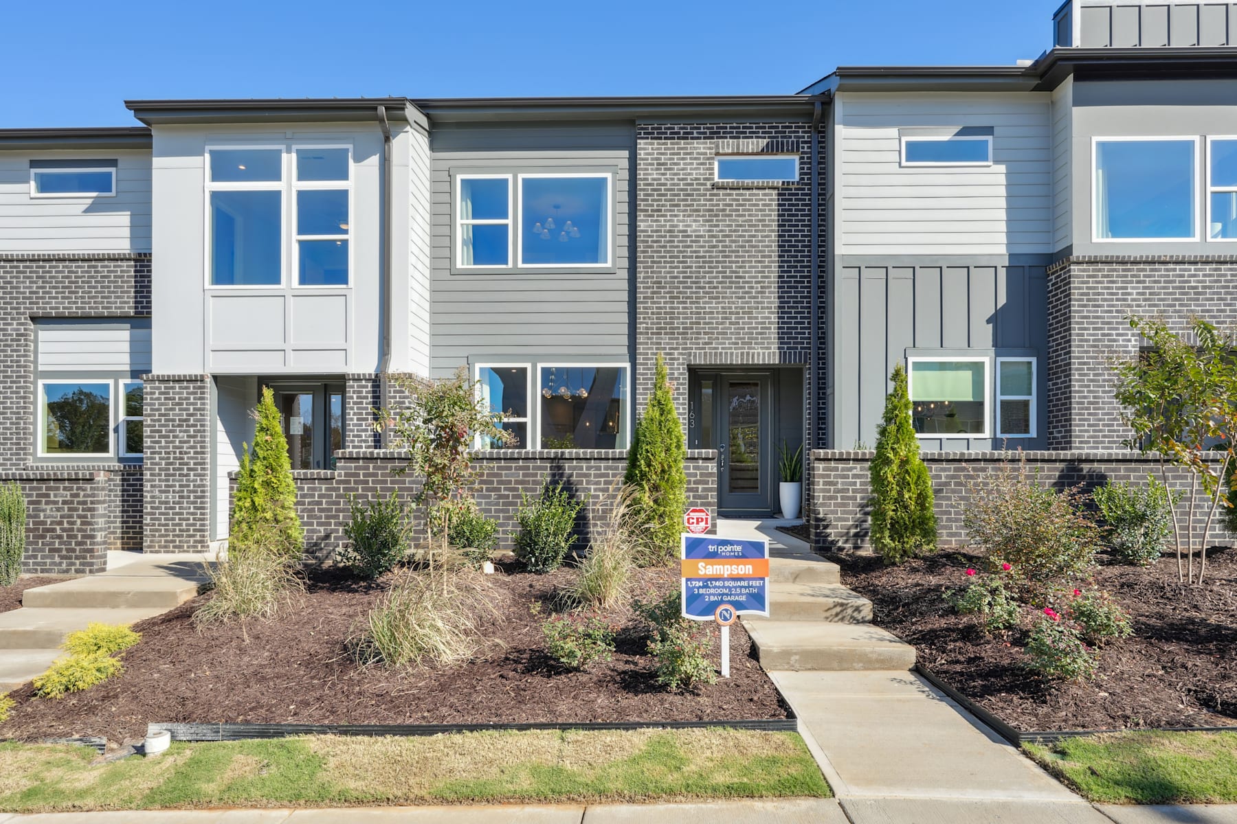 A modern townhouse with a well-landscaped front yard, featuring a brick and siding exterior, large windows, and a paved walkway leading to the entrance.