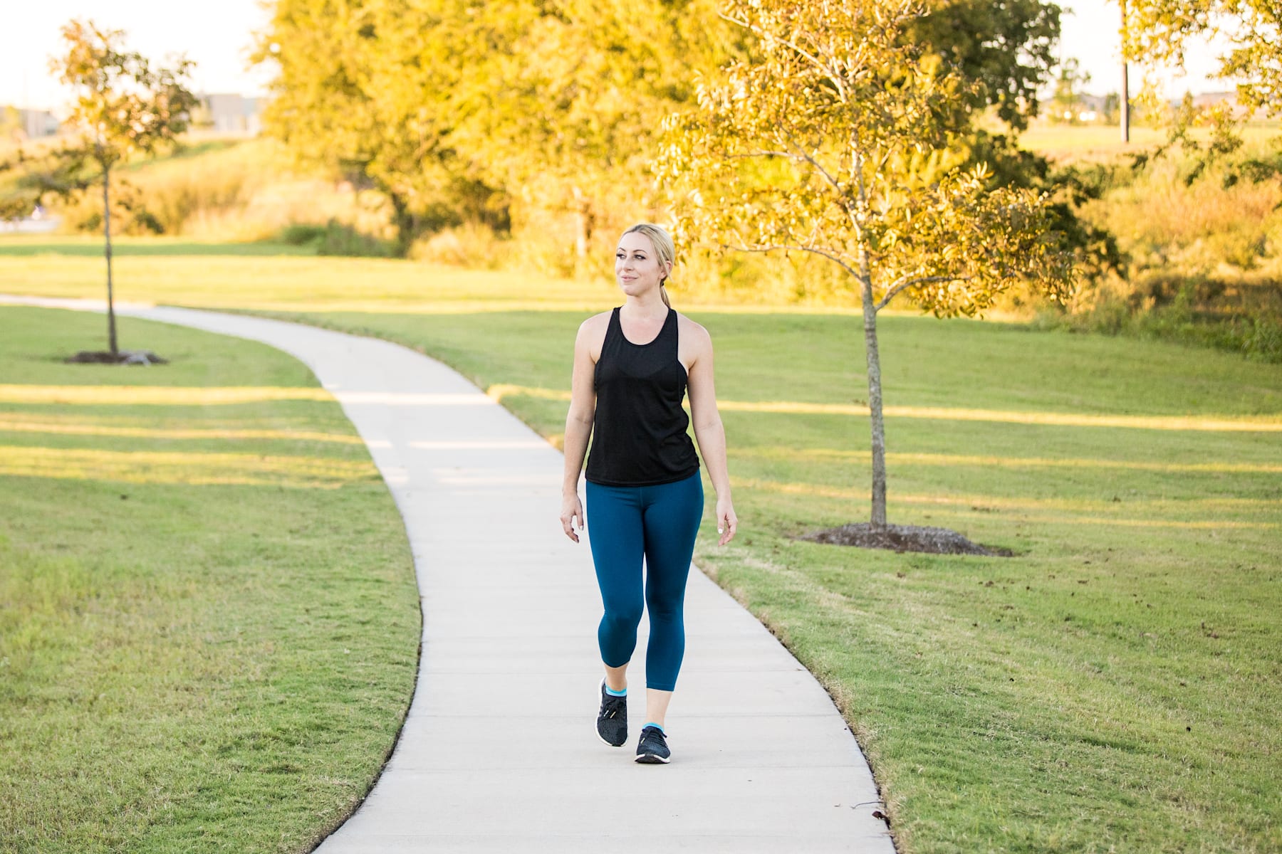 A woman in athletic clothing walks along a paved path surrounded by lush greenery and vibrant yellow trees in a park setting.