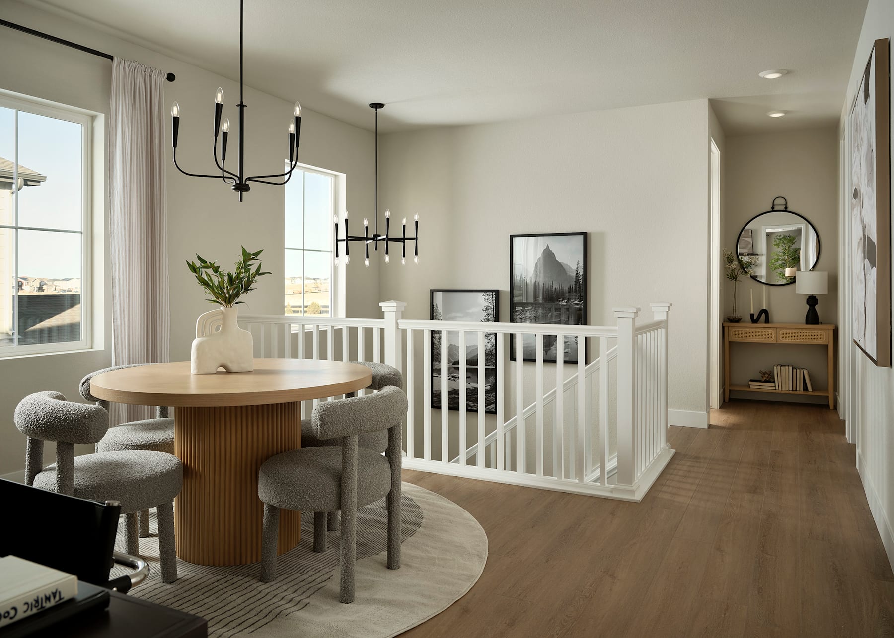 A cozy and well-designed dining area with a round wooden table, upholstered chairs, and a modern chandelier, set against a backdrop of a hallway with a white railing and artwork on the walls.