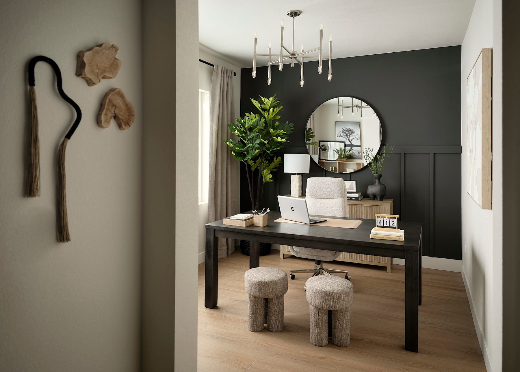 A modern and stylish home office space with a dark-colored accent wall, a wooden desk, and decorative elements such as a round mirror and potted plants.