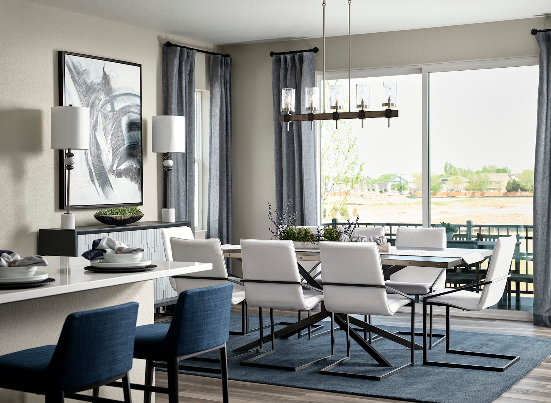 A modern and stylish dining room with a large window overlooking a scenic outdoor area, featuring a rectangular dining table surrounded by white and blue upholstered chairs, a black and white abstract artwork on the wall, and a sleek chandelier overhead.