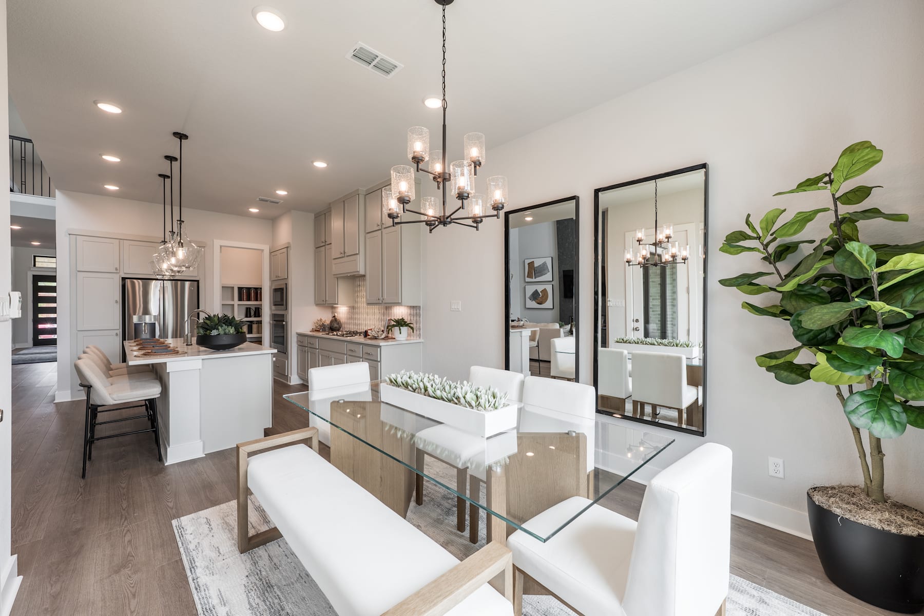 A modern and stylish kitchen and dining area with white cabinets, wooden floors, and a large window providing natural light, complemented by a potted plant and framed artwork on the walls.