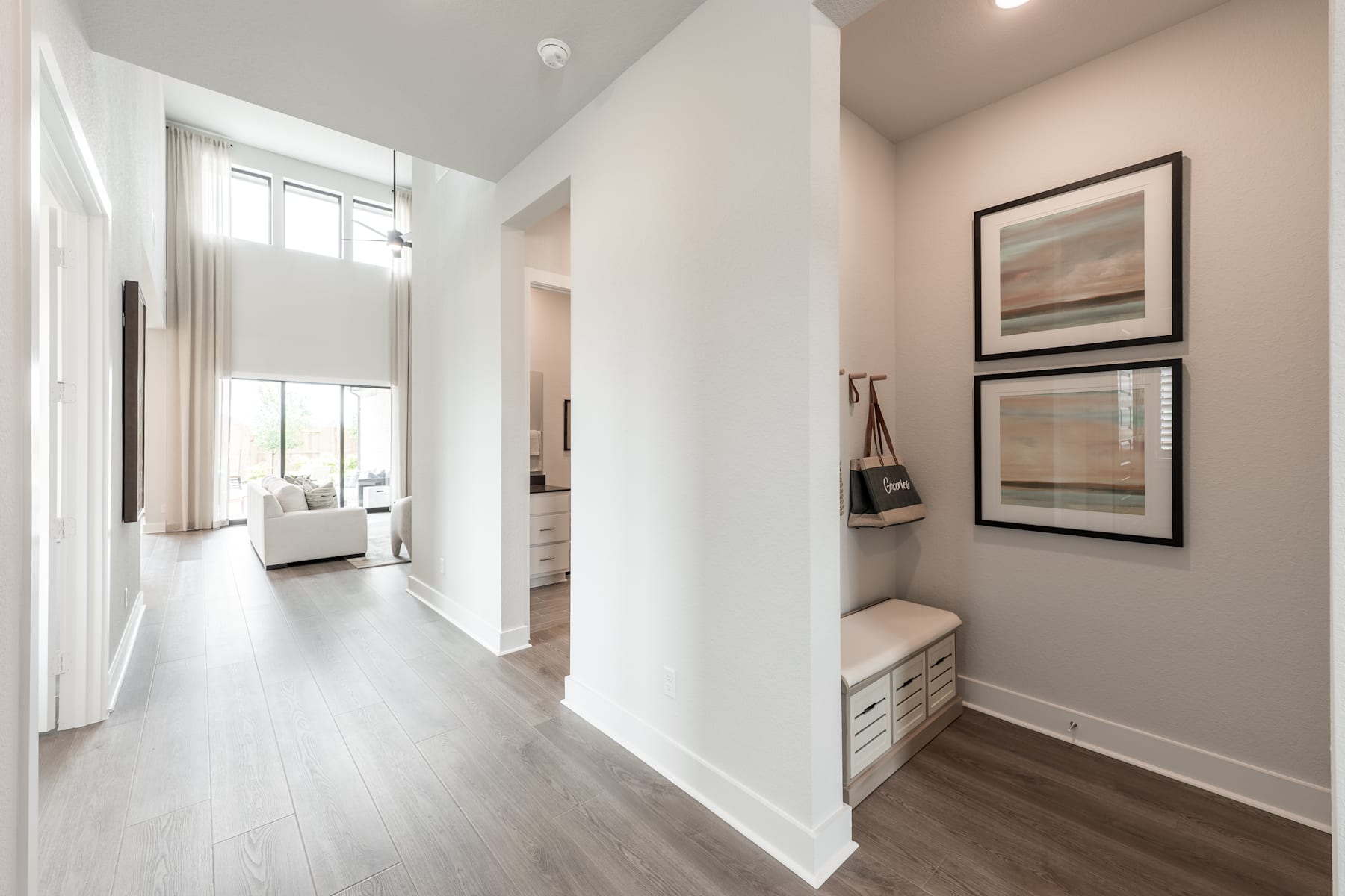 A bright and airy hallway with white walls, hardwood floors, and framed artwork on the walls, leading to a room in the background.