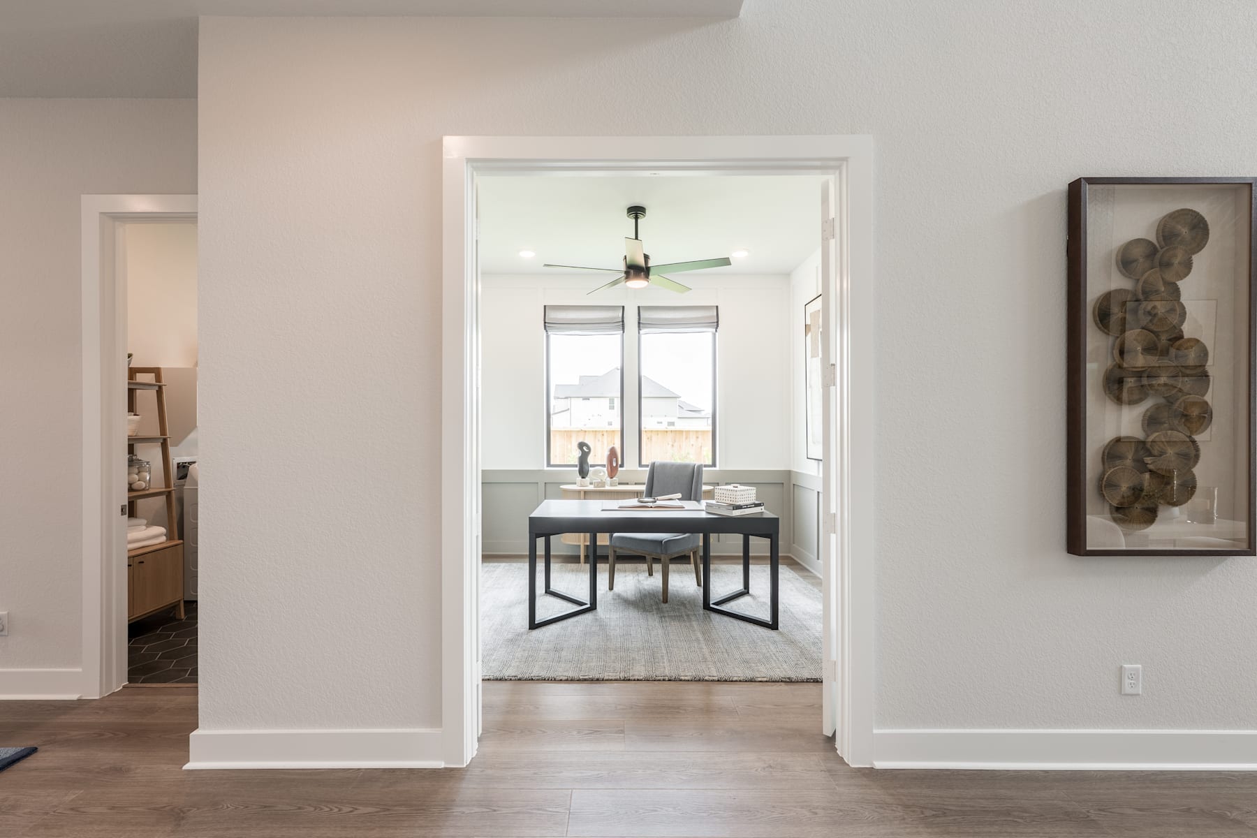 A modern, minimalist home office with a desk, chairs, and artwork on the walls, visible through an open doorway.