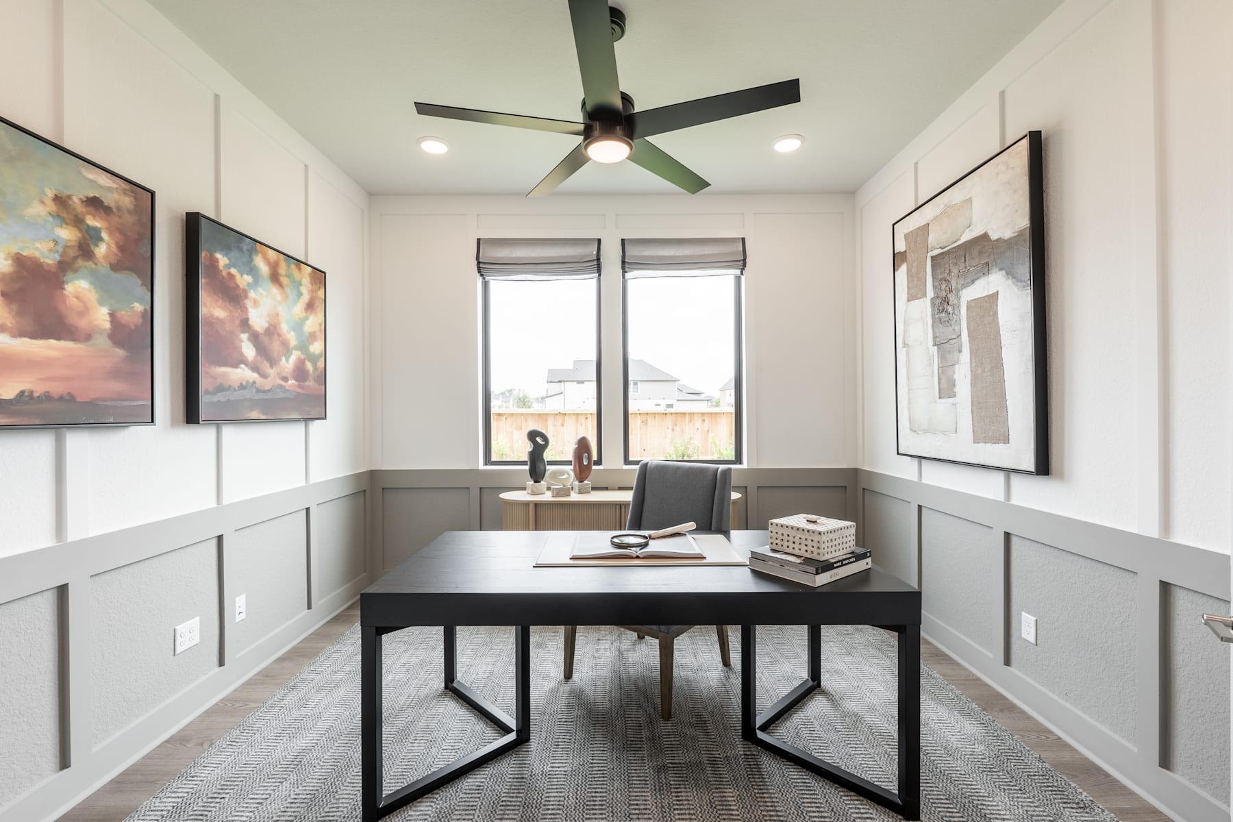 A modern and minimalist home office space with a large desk, a ceiling fan, and artwork on the walls, set against a bright and airy background.