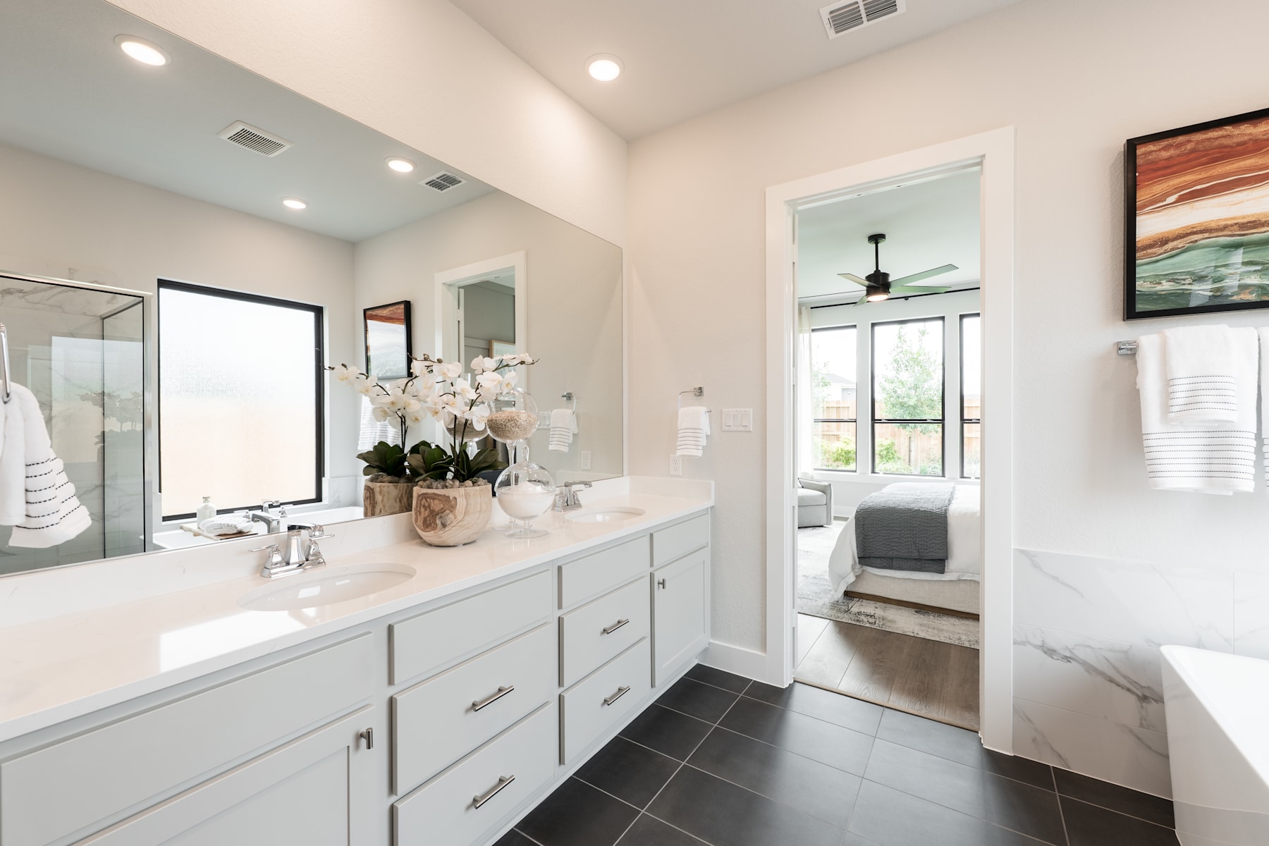 A modern and spacious bathroom with white cabinets, a large vanity mirror, and a view of an adjacent bedroom through an open doorway.