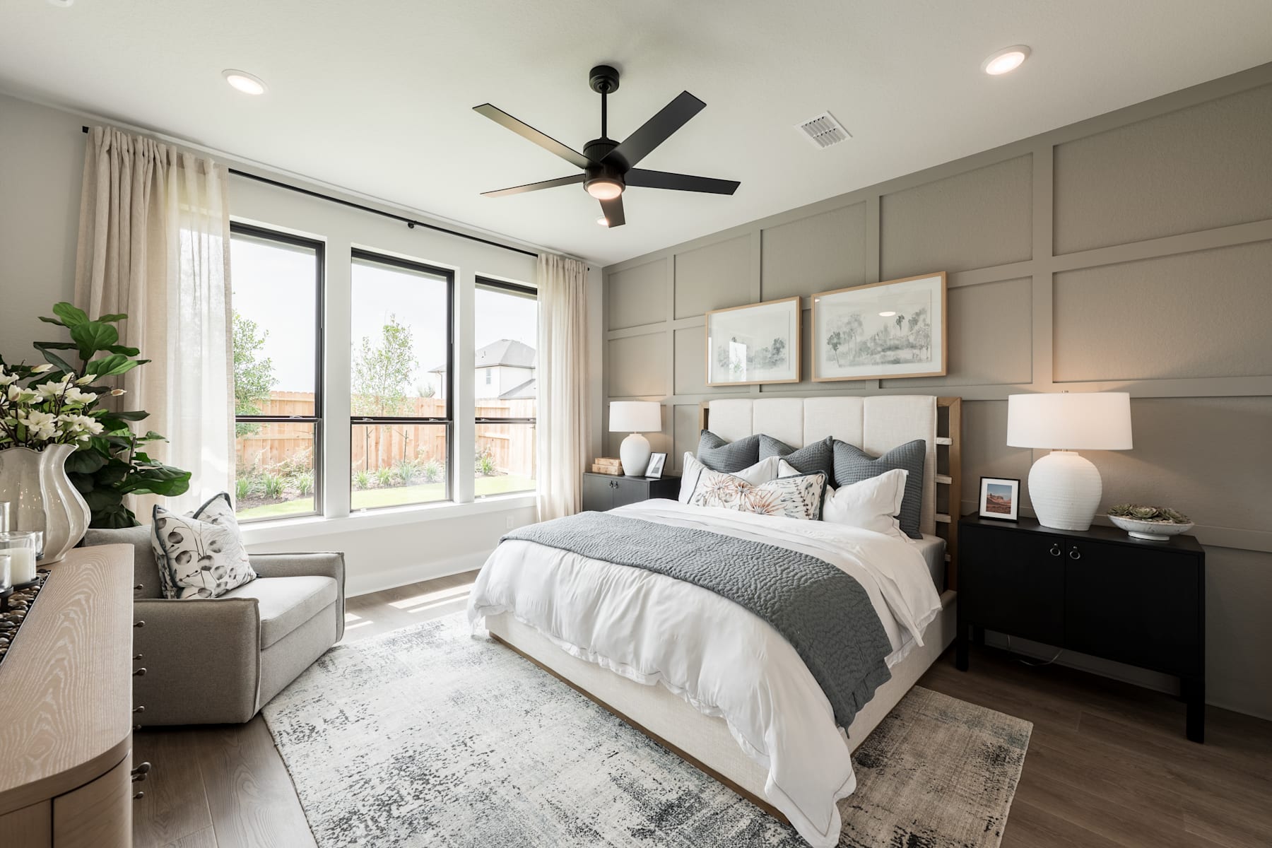 A cozy and well-designed bedroom with a large bed, neutral-toned decor, and a ceiling fan, surrounded by large windows that allow natural light to fill the space.