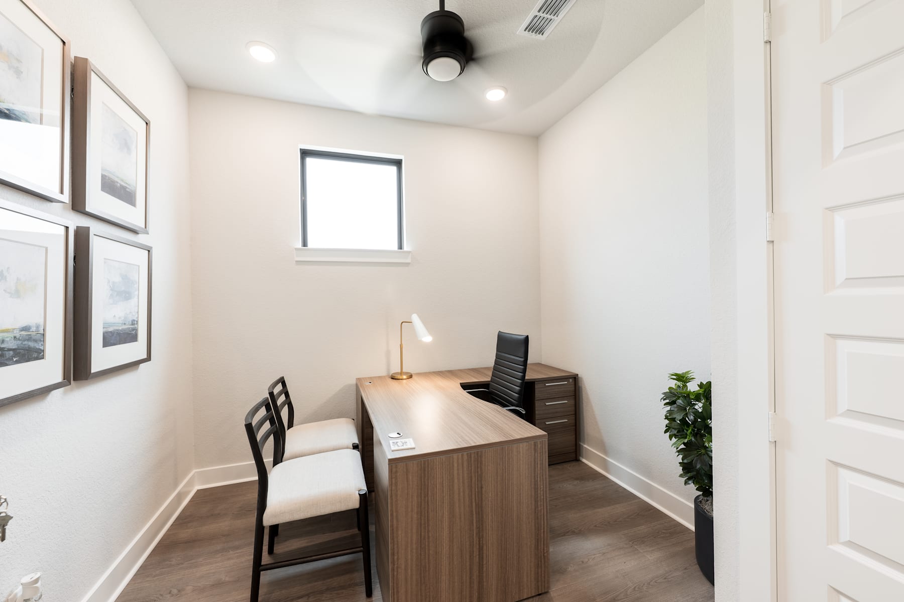 A small, well-lit office space with a wooden desk, a chair, and framed artwork on the walls.