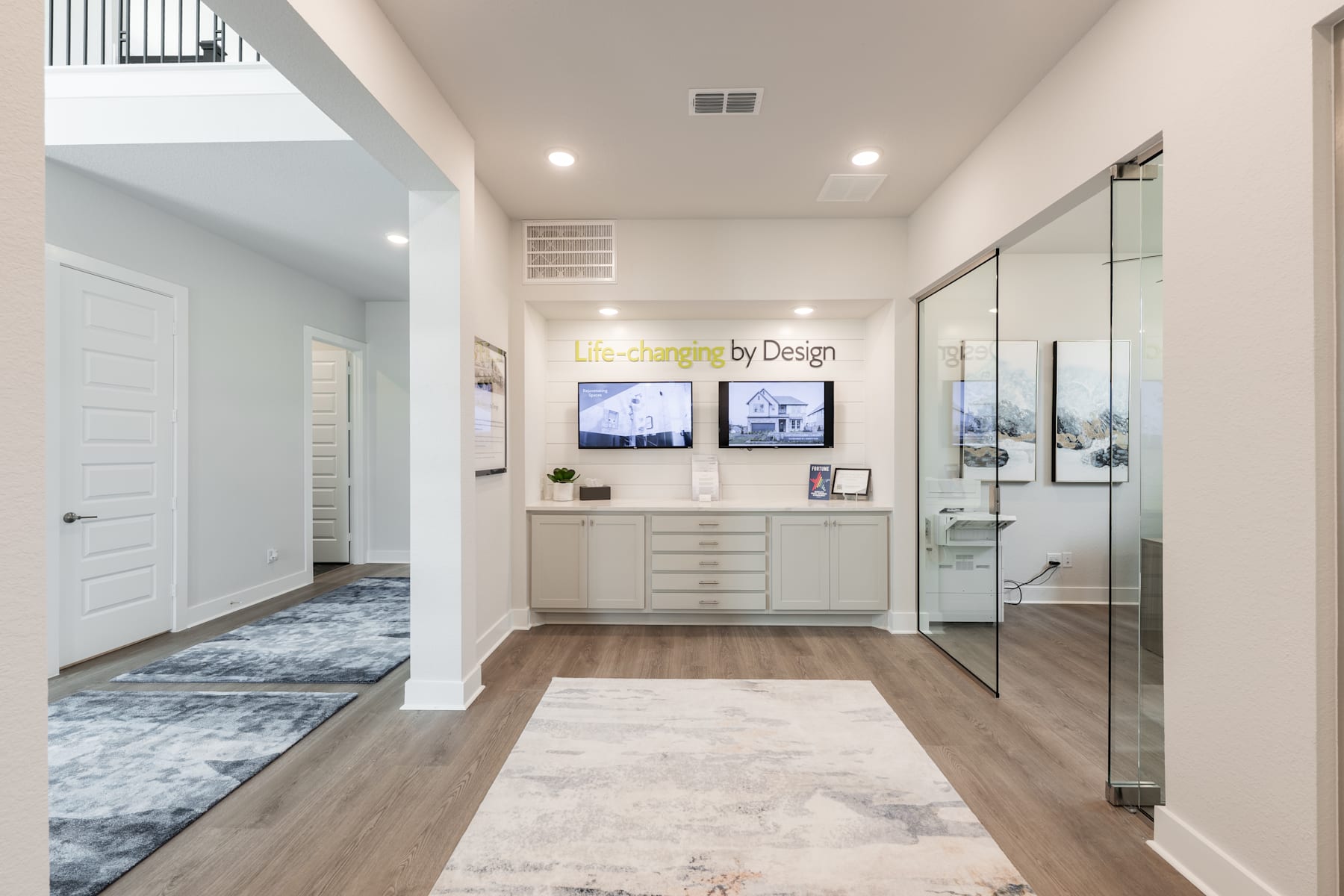 A spacious and modern entryway with a sleek white cabinet, multiple screens, and a patterned rug leading into a well-lit hallway.