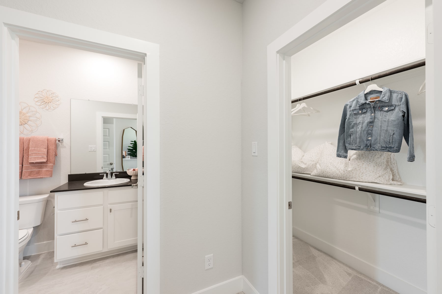 A bright and airy bathroom with a vanity and mirror, leading into a closet area with a hanging denim jacket.