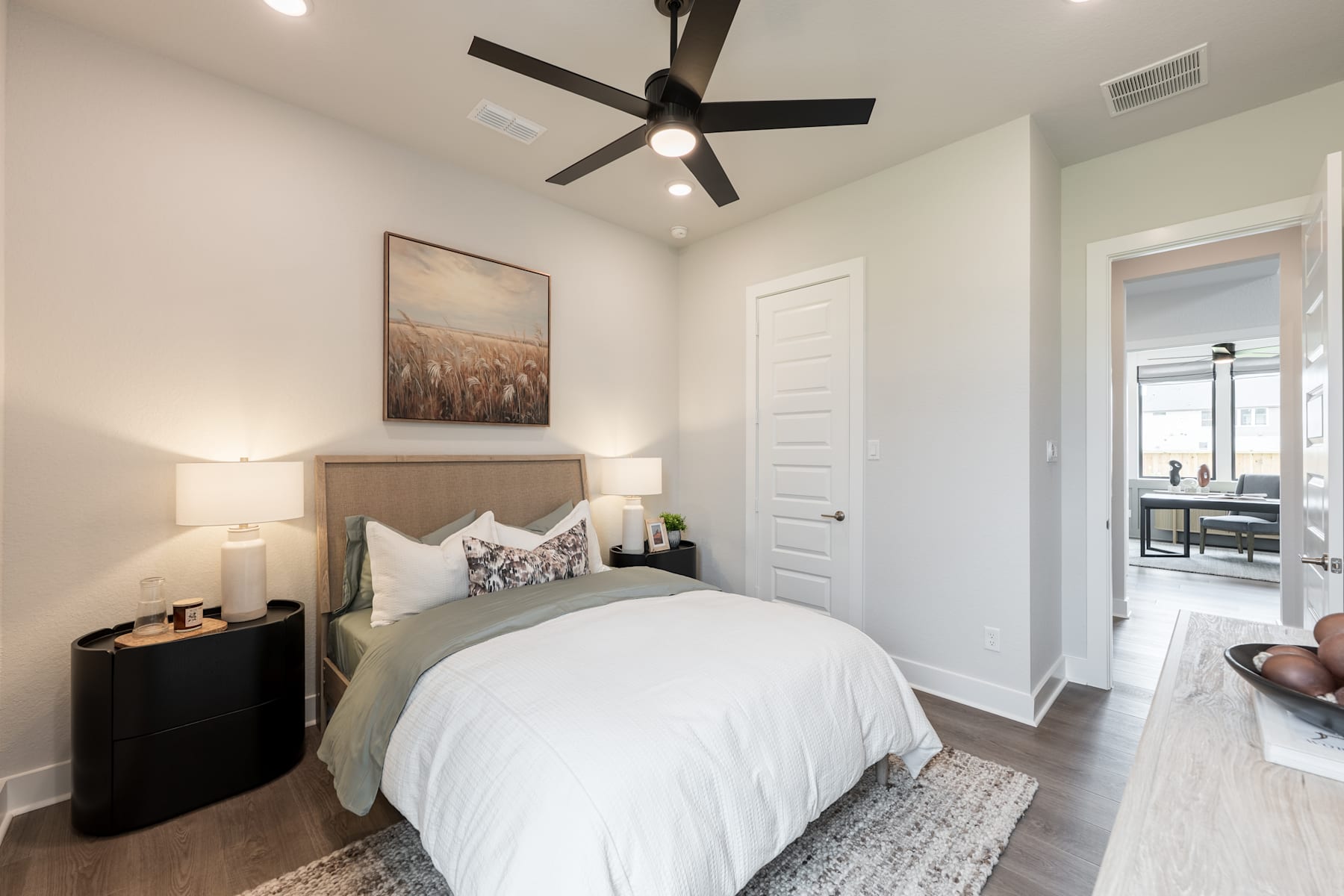 A cozy and well-furnished bedroom with a large bed, a ceiling fan, and a framed artwork on the wall, along with a dresser and a door leading to an adjacent room.