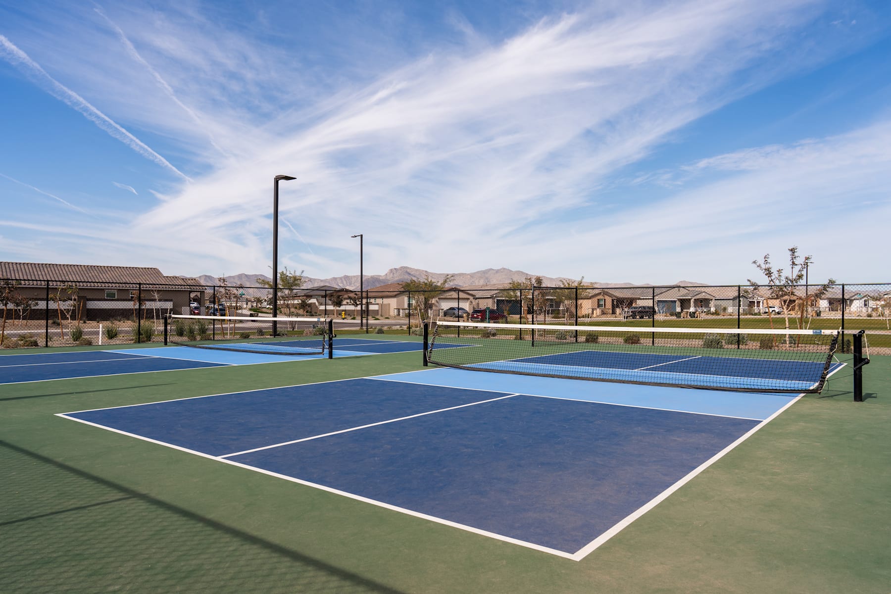 The image shows a tennis court surrounded by residential buildings and a mountainous landscape in the background, with a clear blue sky overhead.