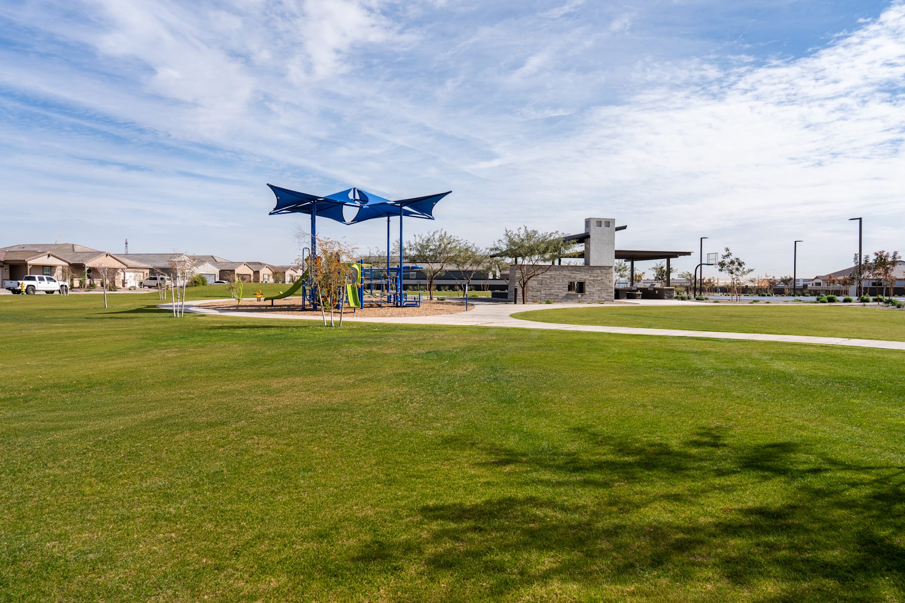 A grassy park with playground equipment, surrounded by residential buildings and a blue-and-white shade structure in the background.