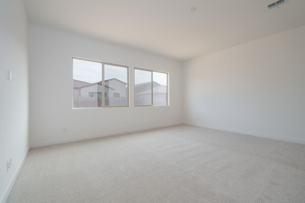 An empty, bright room with white walls, a tiled floor, and several windows overlooking residential buildings in the background.