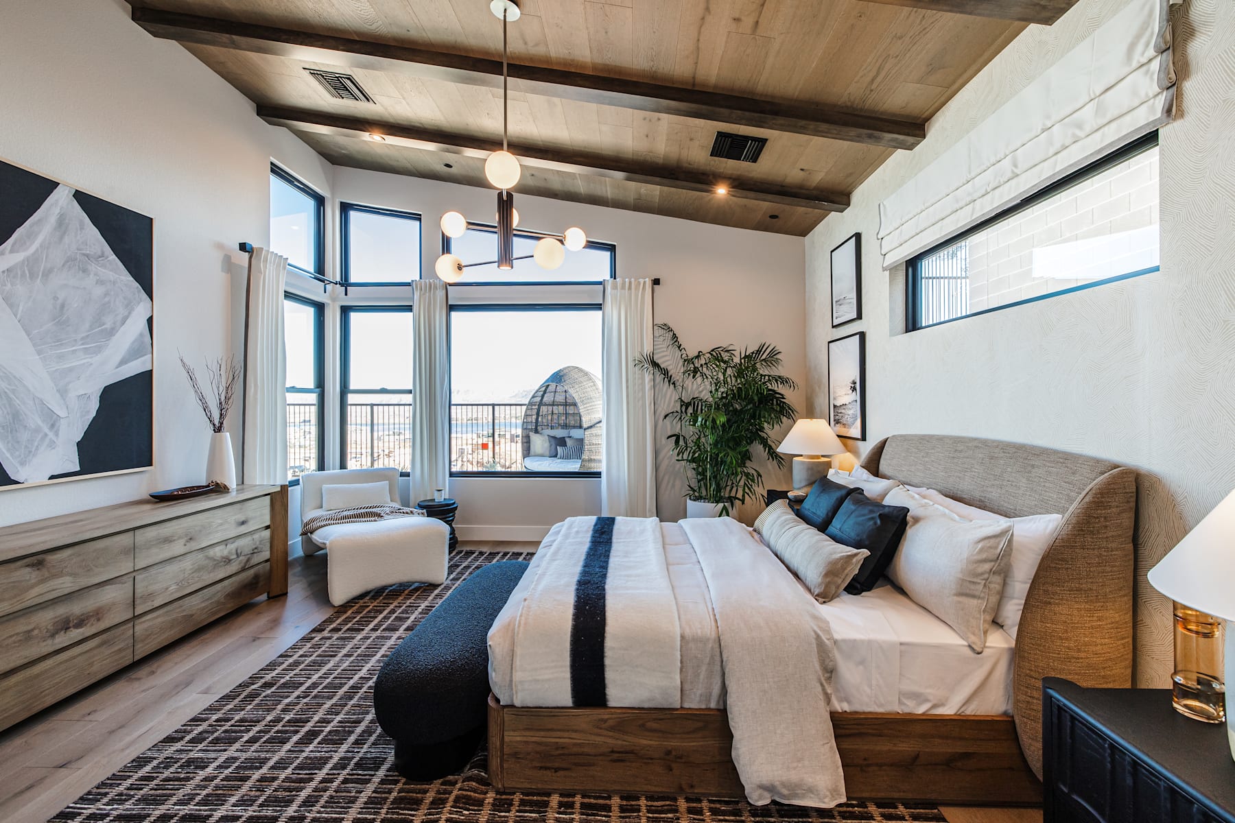 A cozy and inviting bedroom with a wooden ceiling, large windows, and a plush bed adorned with neutral-toned bedding, complemented by a dresser and other furnishings.