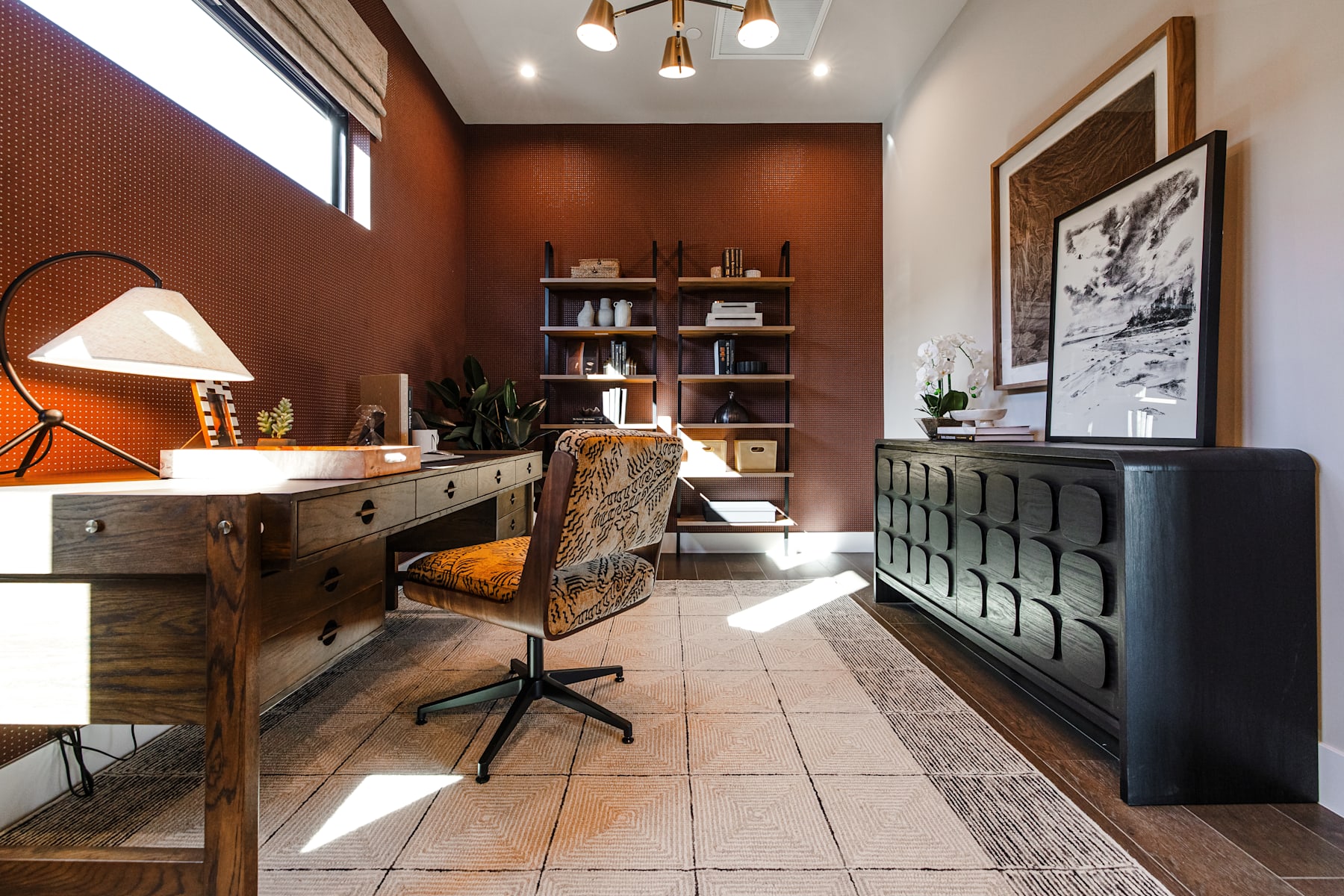 A cozy and well-designed home office space with a wooden desk, shelves, and a black filing cabinet, set against a warm-toned wall and accented by modern lighting fixtures.