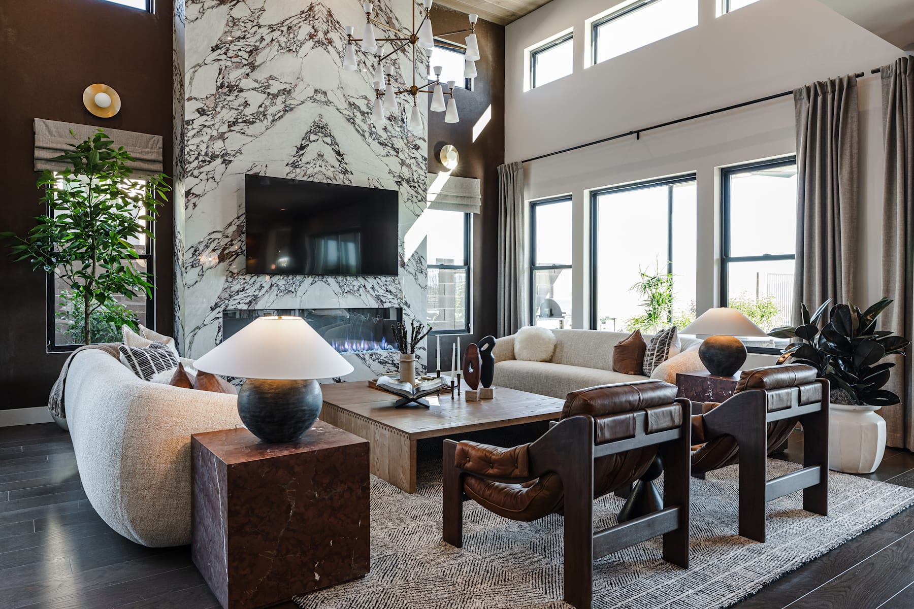 A spacious and modern living room with a large marble fireplace, plush seating, and a wooden dining table surrounded by chairs, all set against a backdrop of large windows and lush greenery.