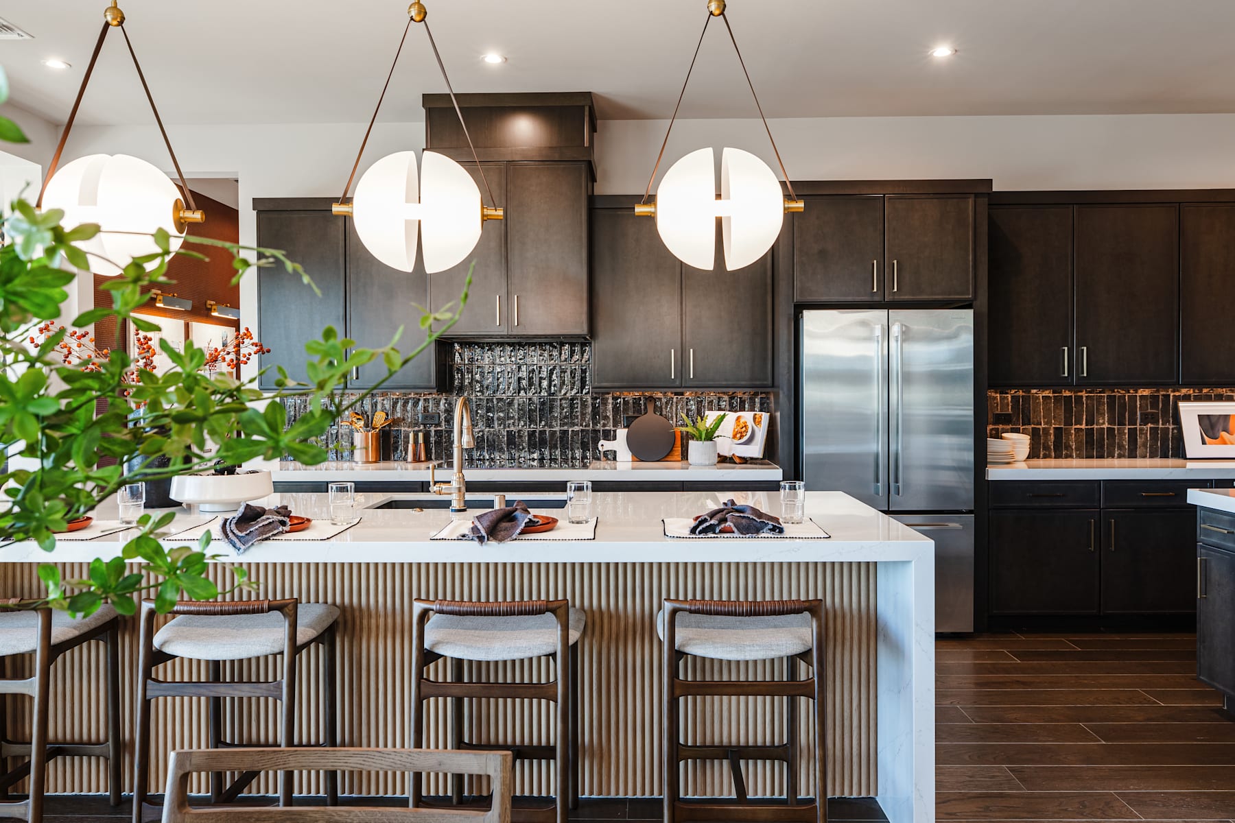 A modern and stylish kitchen with a large island, dark cabinets, and pendant lights, surrounded by potted plants and a cozy atmosphere.