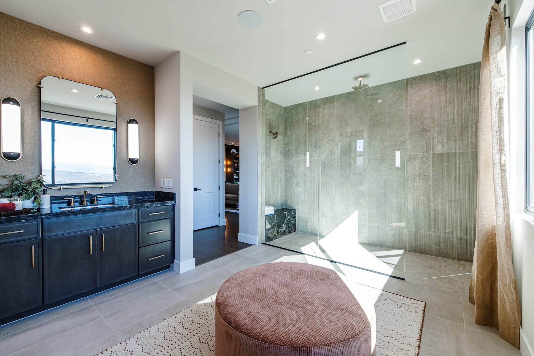 A modern bathroom with a large glass shower enclosure, dark cabinets, and a plush pink ottoman in the foreground.