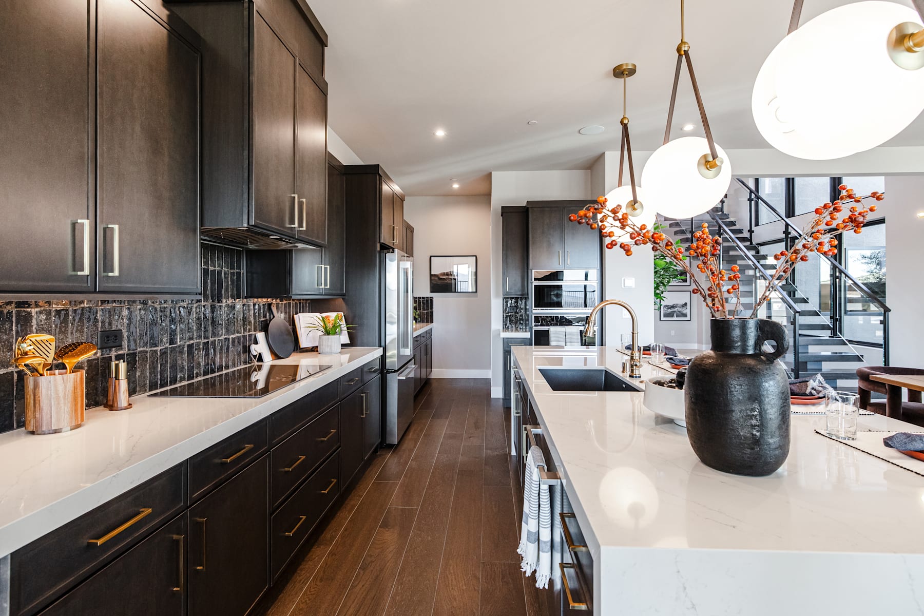 A modern and stylish kitchen with dark cabinets, a white countertop, and decorative elements such as a vase with flowers and pendant lighting fixtures.