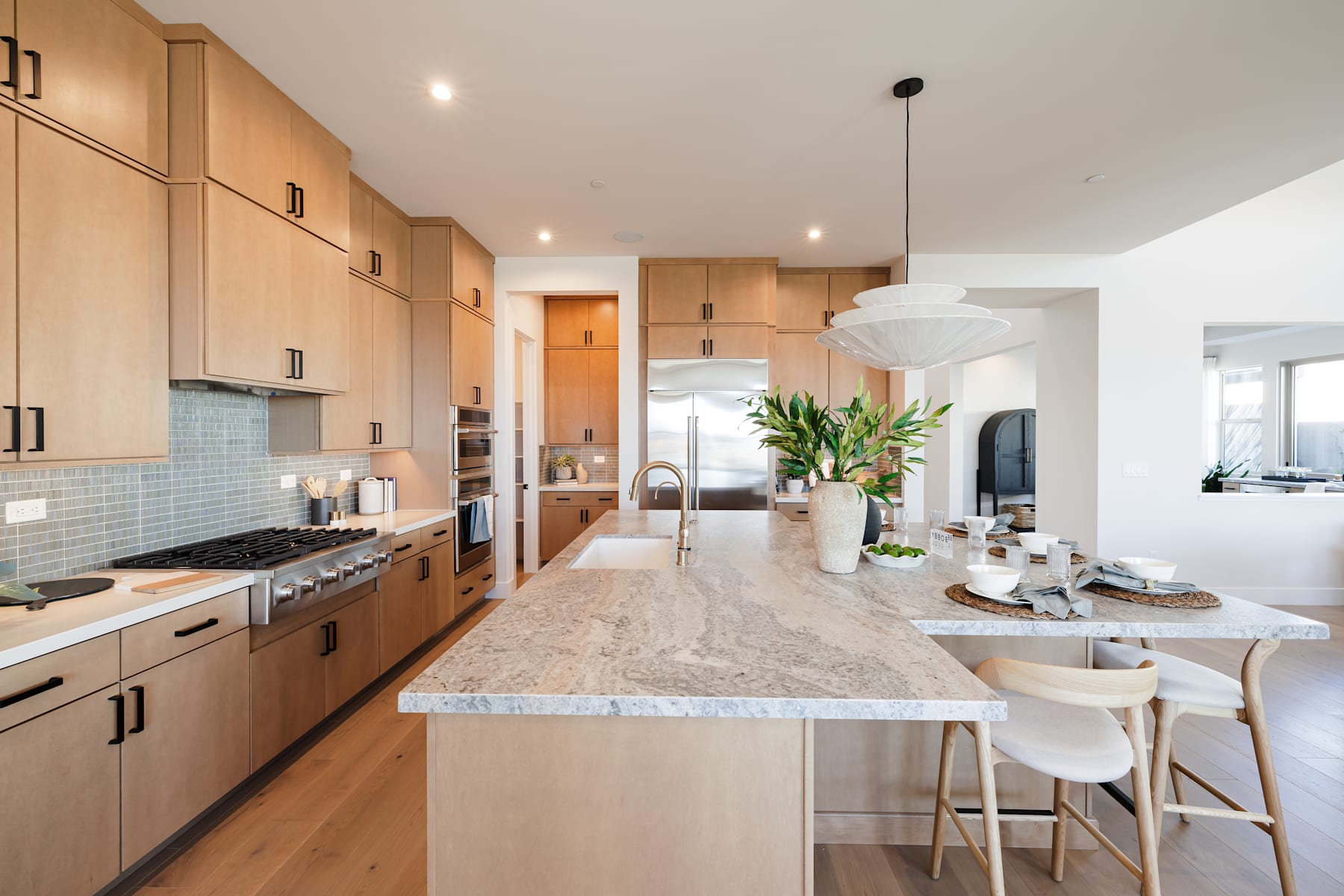 A modern, open-concept kitchen with light wood cabinetry, a large marble-topped island, and a dining area with white chairs in the background.