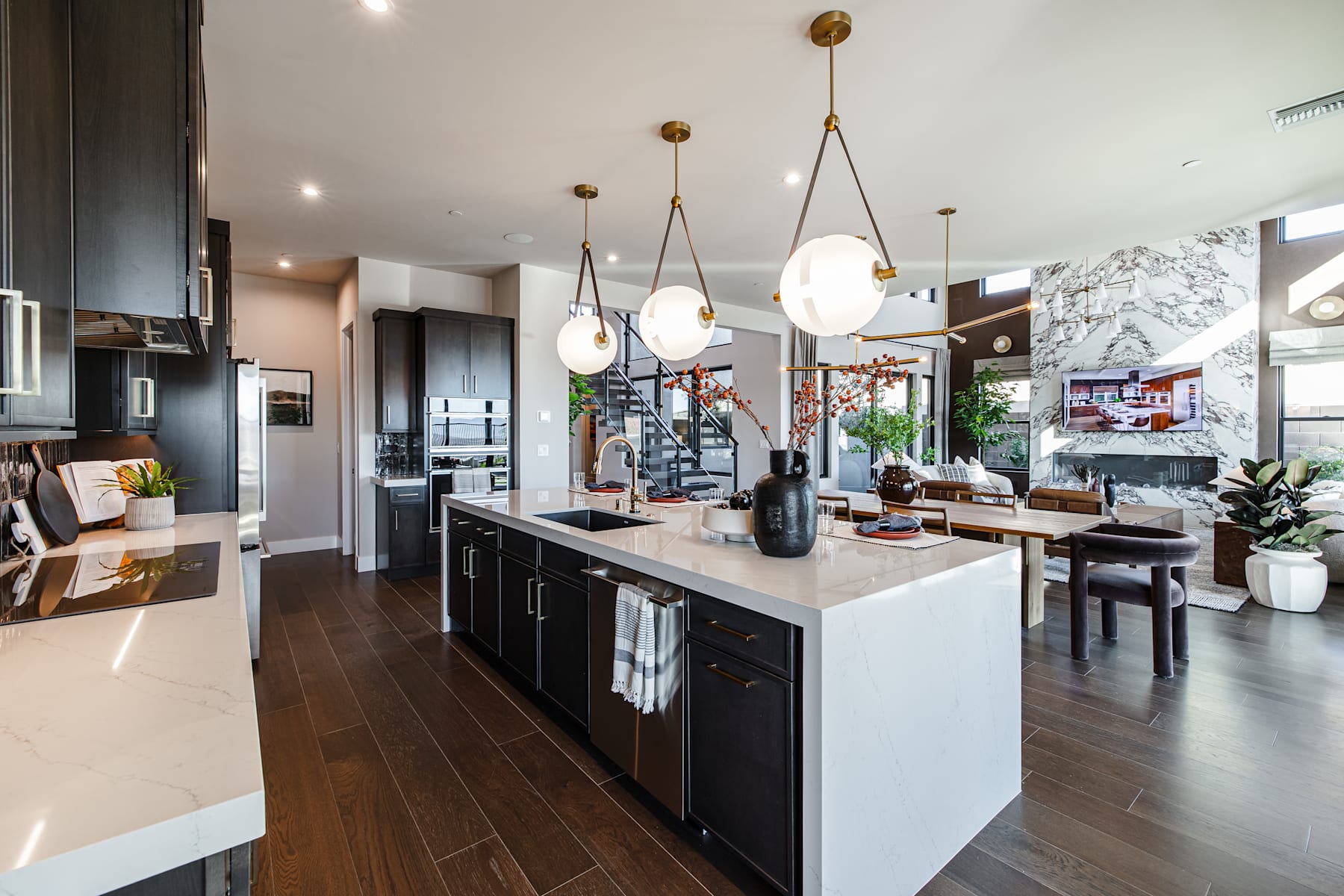 A modern and spacious kitchen with a large island, dark cabinets, and pendant lighting fixtures, set against a backdrop of a dining area and living room.