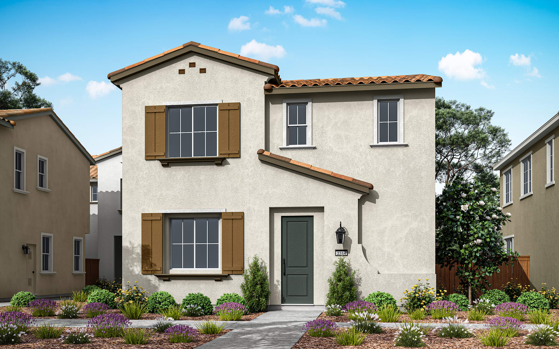 A two-story Mediterranean-style house with a tiled roof, surrounded by a well-landscaped garden featuring various flowering plants and shrubs, set against a clear blue sky with scattered clouds.