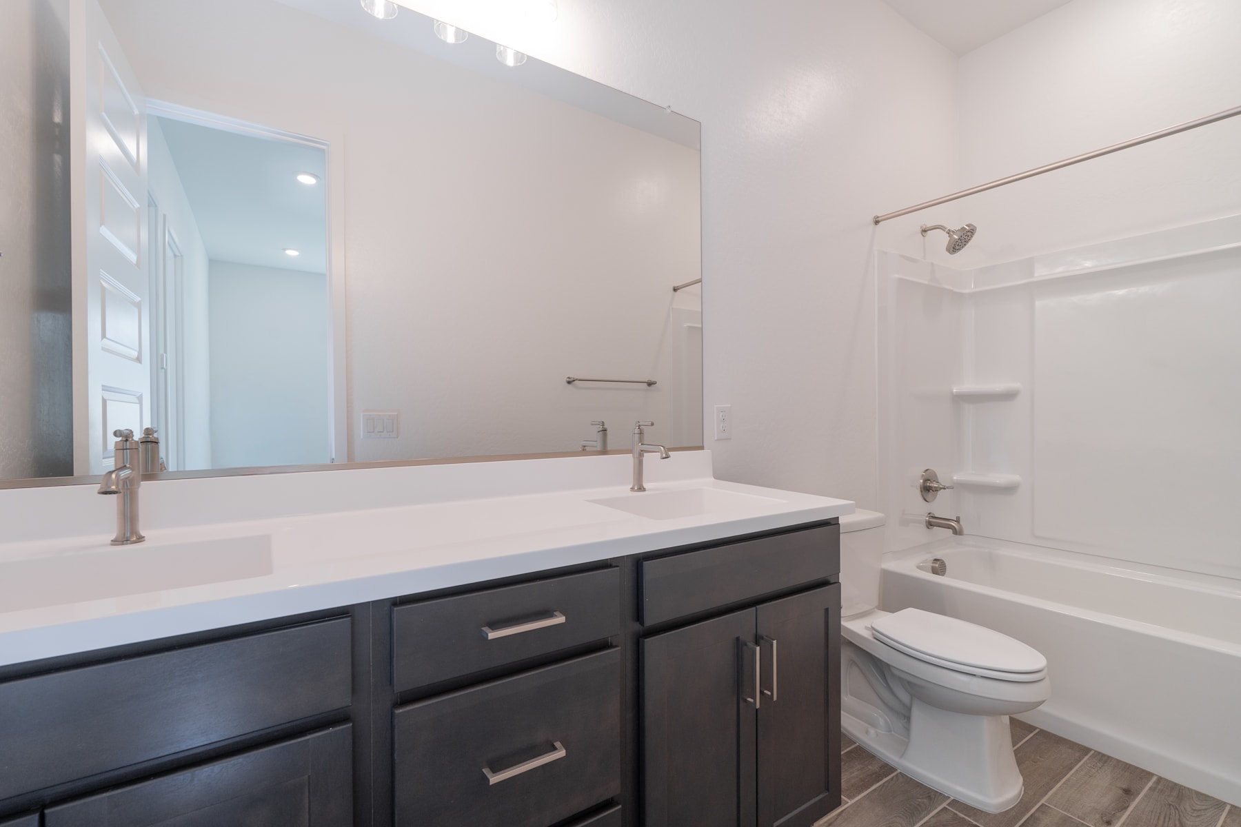 A modern bathroom with a dark vanity cabinet, a large mirror, and a bathtub visible in the background.