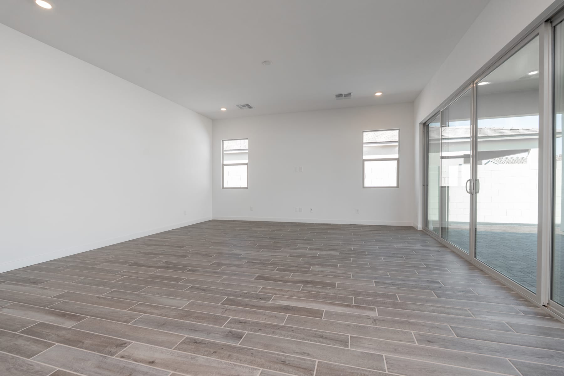 A spacious, minimalist room with white walls, large windows, and a wooden-tiled floor.