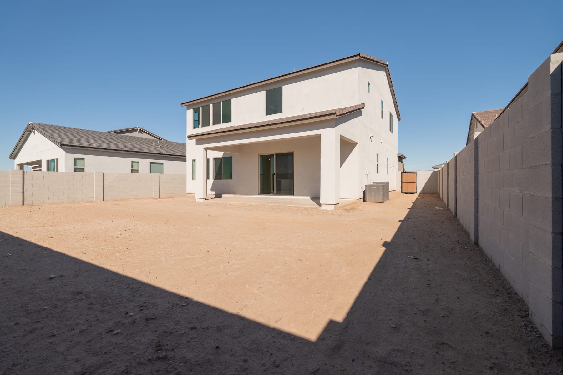 A modern two-story residential building with a paved driveway and a dirt-covered yard in the foreground, set against a clear blue sky.