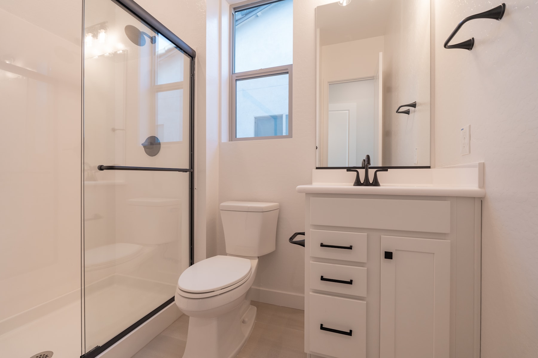 A modern and minimalist bathroom with a white vanity, toilet, and glass shower enclosure, set against a bright and airy backdrop.