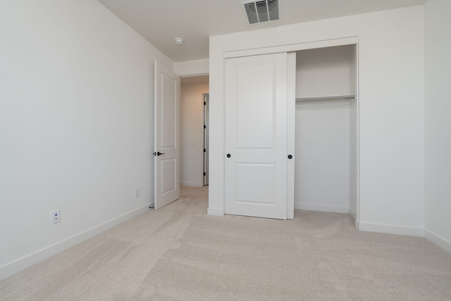 A simple, minimalist bedroom with white walls, a closet, and a door leading to another room.