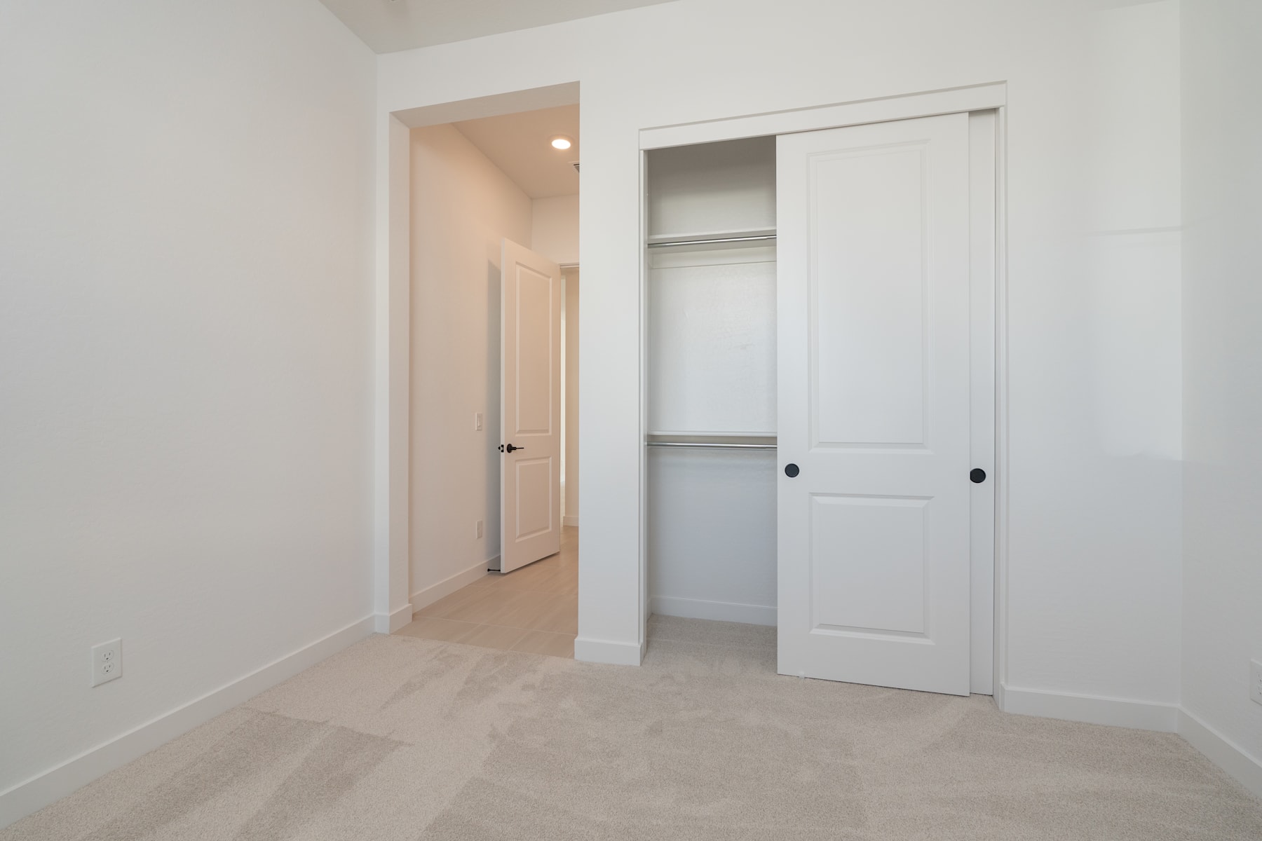 A simple, minimalist bedroom with a closet door and a hallway visible in the background, all in a clean, white color scheme.